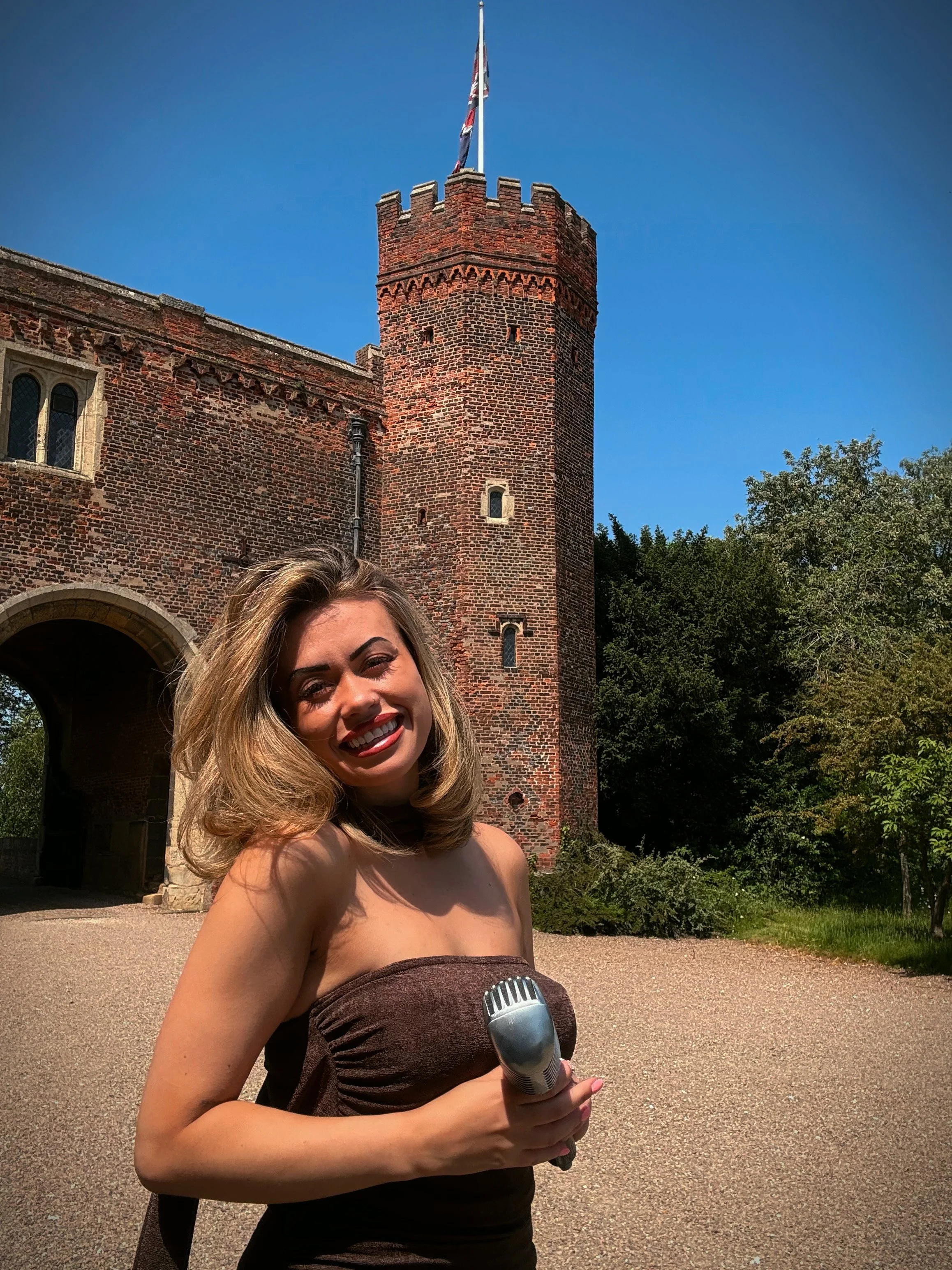 A woman with blonde hair smiling, holding a microphone, standing in front of a red brick castle tower with a flag on top and a clear blue sky.