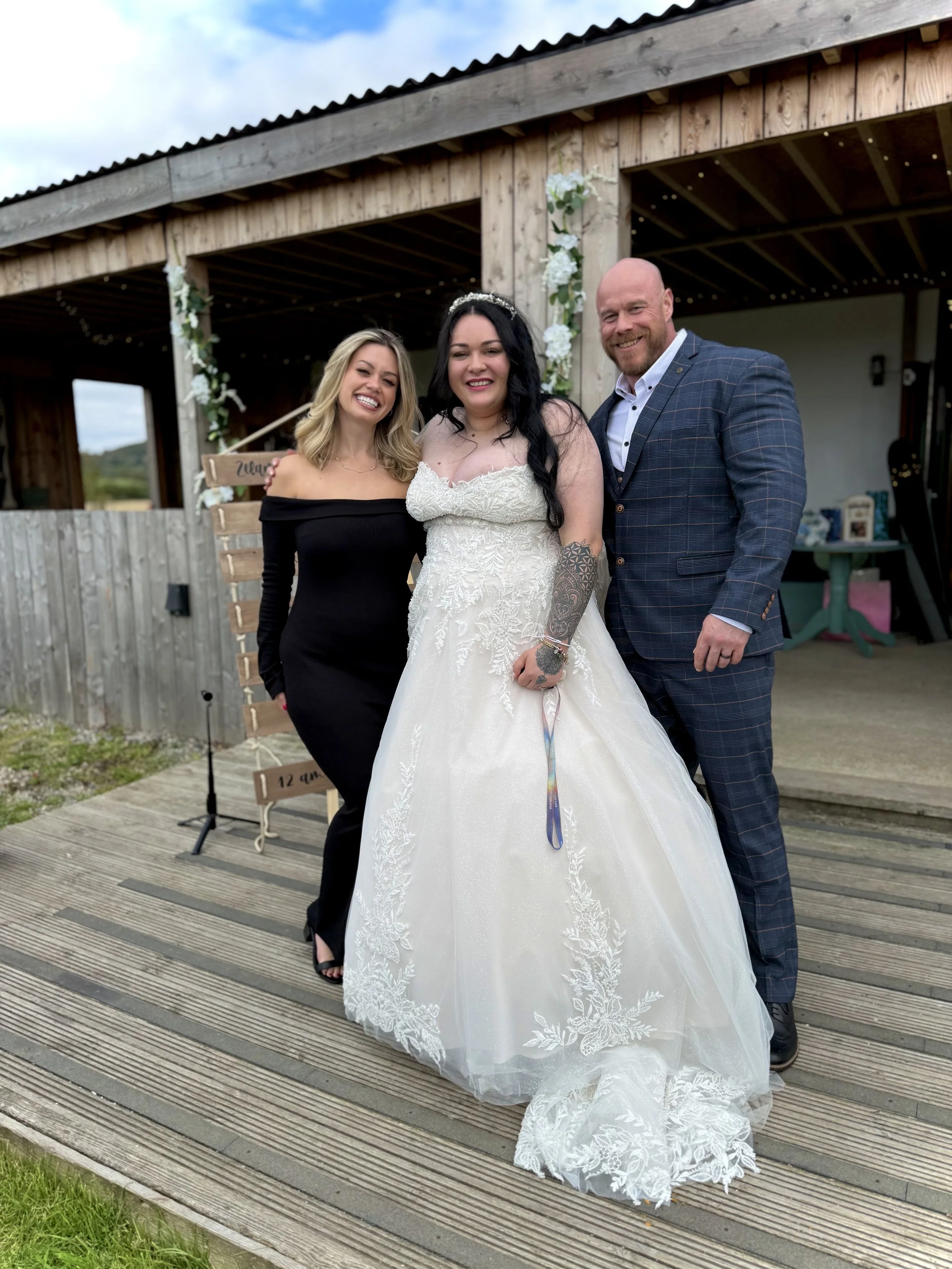 Three people, two women and one man, standing together at a wedding venue outdoors, with a wooden barn in the background. The woman in the center is wearing a wedding dress, and all are smiling for the photo.