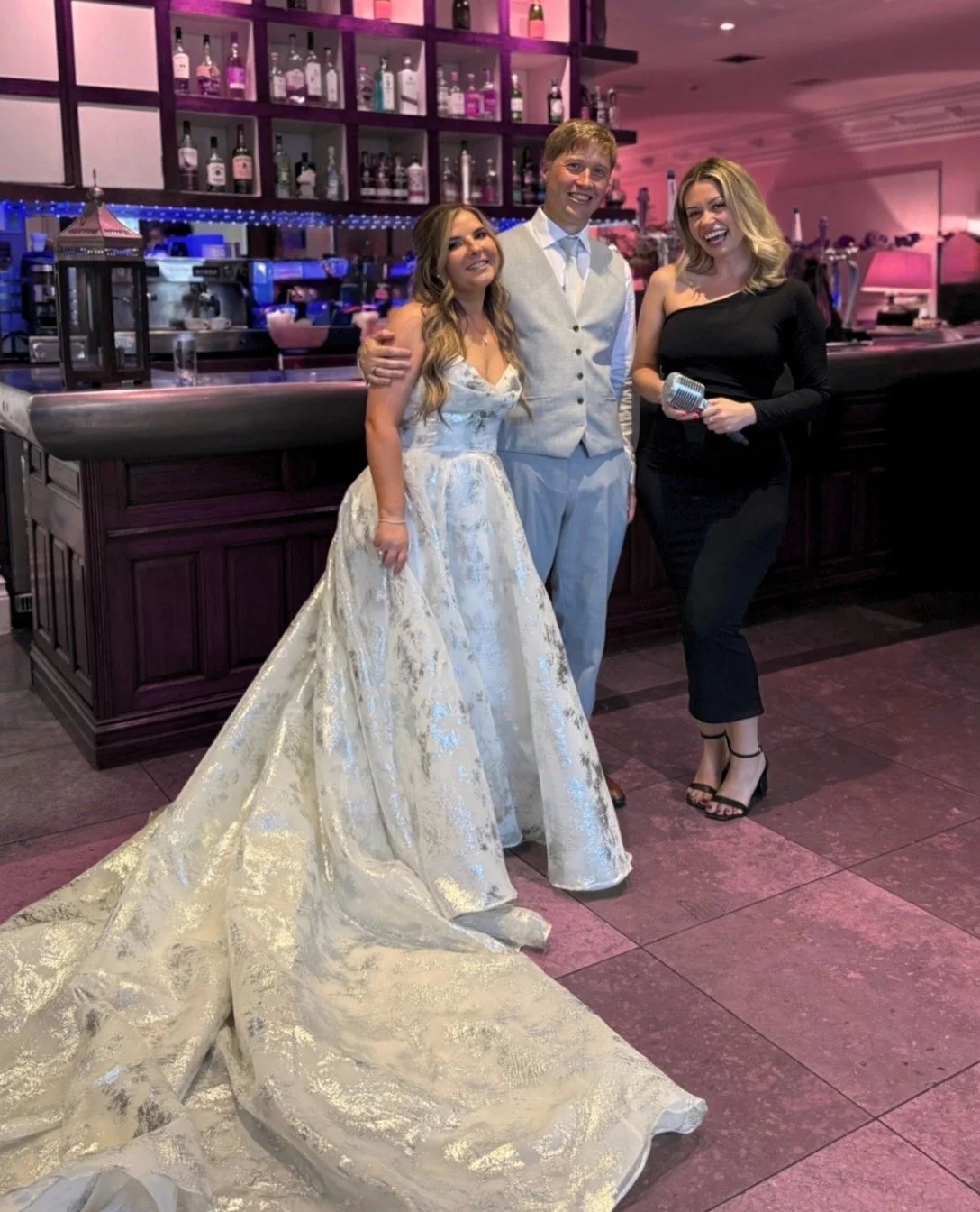 A bride in a white and gold gown, a groom in a tuxedo, and a woman in a black dress holding a microphone are standing together at a bar in a brightly lit reception area, smiling at the camera.