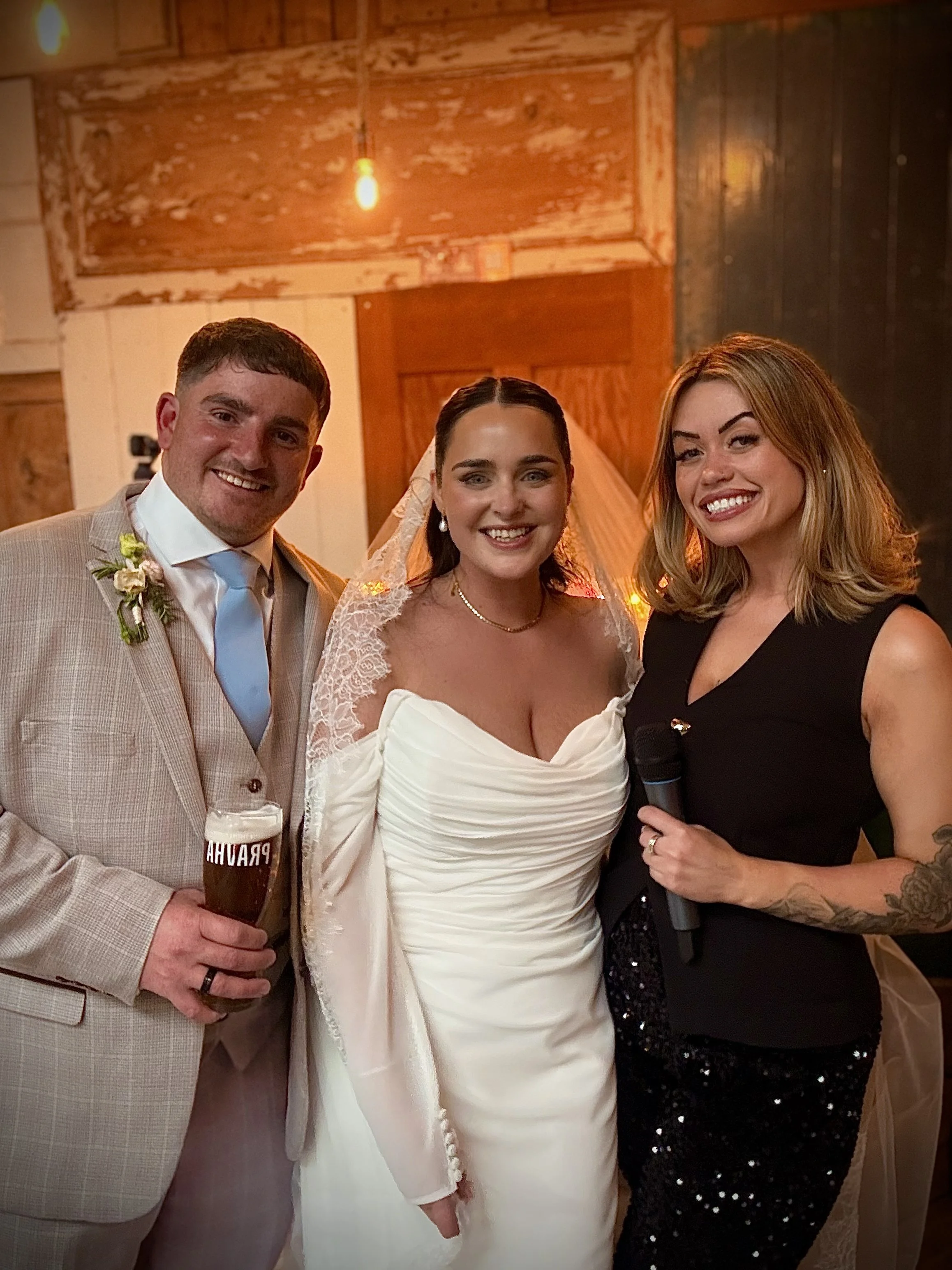 A group of three people dressed formally, celebrating at a wedding reception. The man on the left is wearing a light-colored suit with a blue tie and holding a beer. The woman in the middle is wearing a white wedding dress with lace details and a veil. The woman on the right is wearing a black dress with sequins, holding a microphone, and has tattoos on her right arm. They are all smiling and posing together indoors with warm lighting and wooden decor.