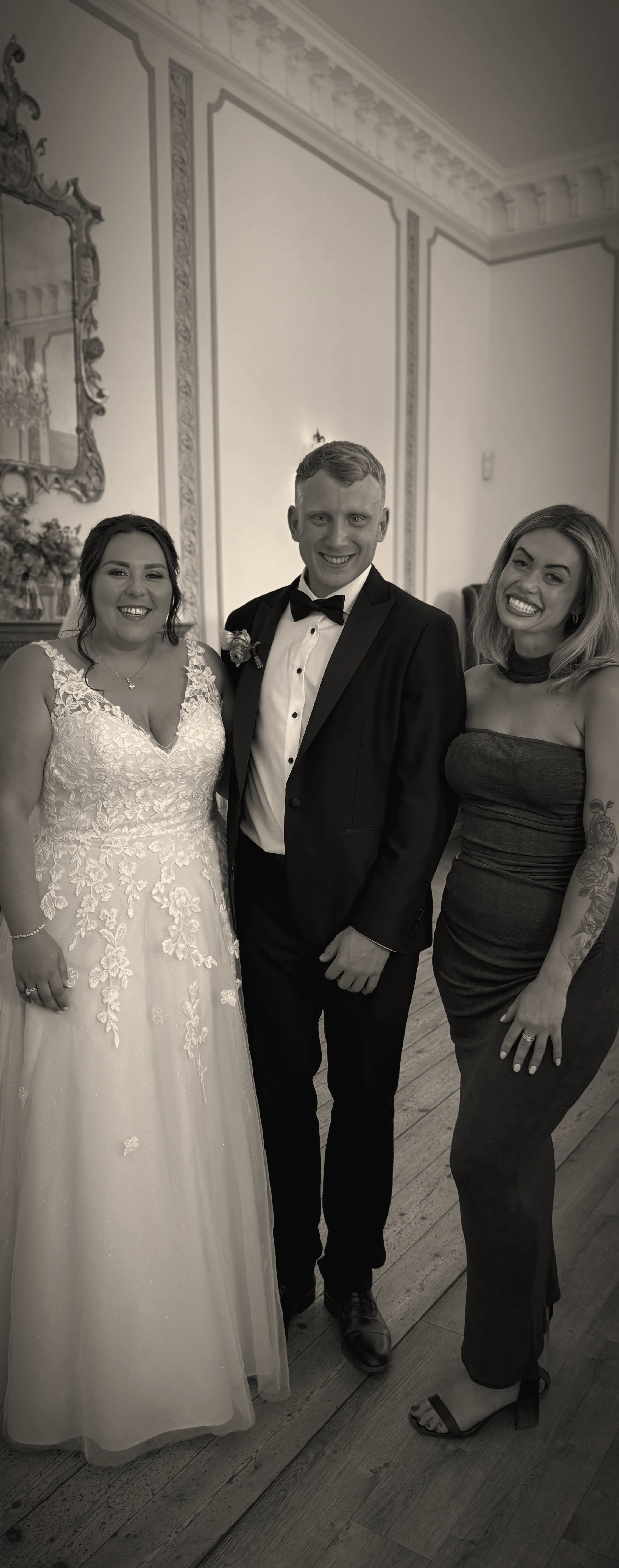 Three people at a formal event, standing in elegant room with ornate decor. The woman on the left is wearing a lace wedding dress, the man in the middle is in a tuxedo, and the woman on the right is in a strapless black dress with tattoos on her arm. All are smiling.