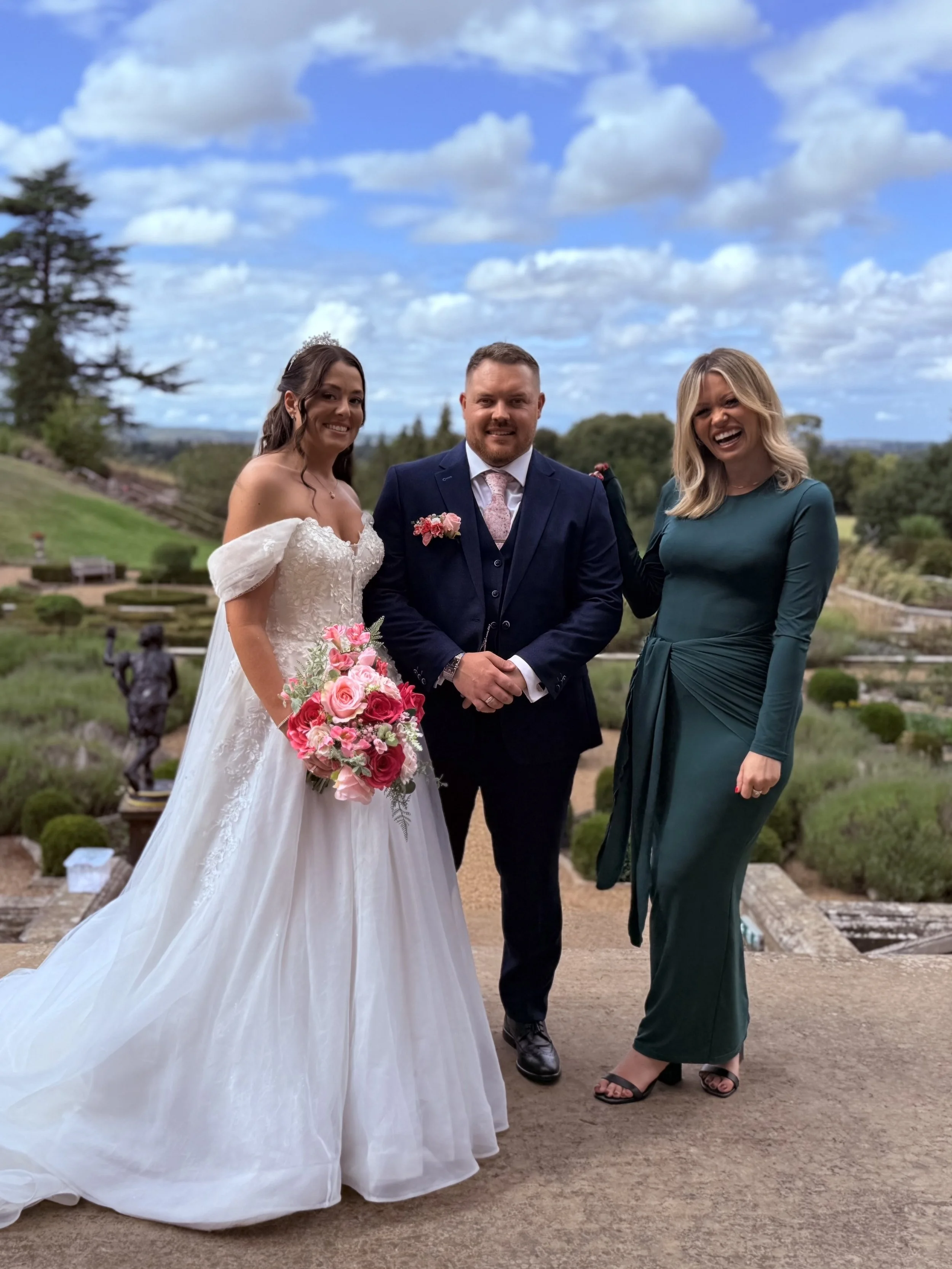 A bride in a white wedding dress holding a bouquet of pink flowers, standing next to a groom in a navy suit, with a woman in an emerald green dress standing to the right, outdoors with a cloudy sky and landscaped garden in the background.