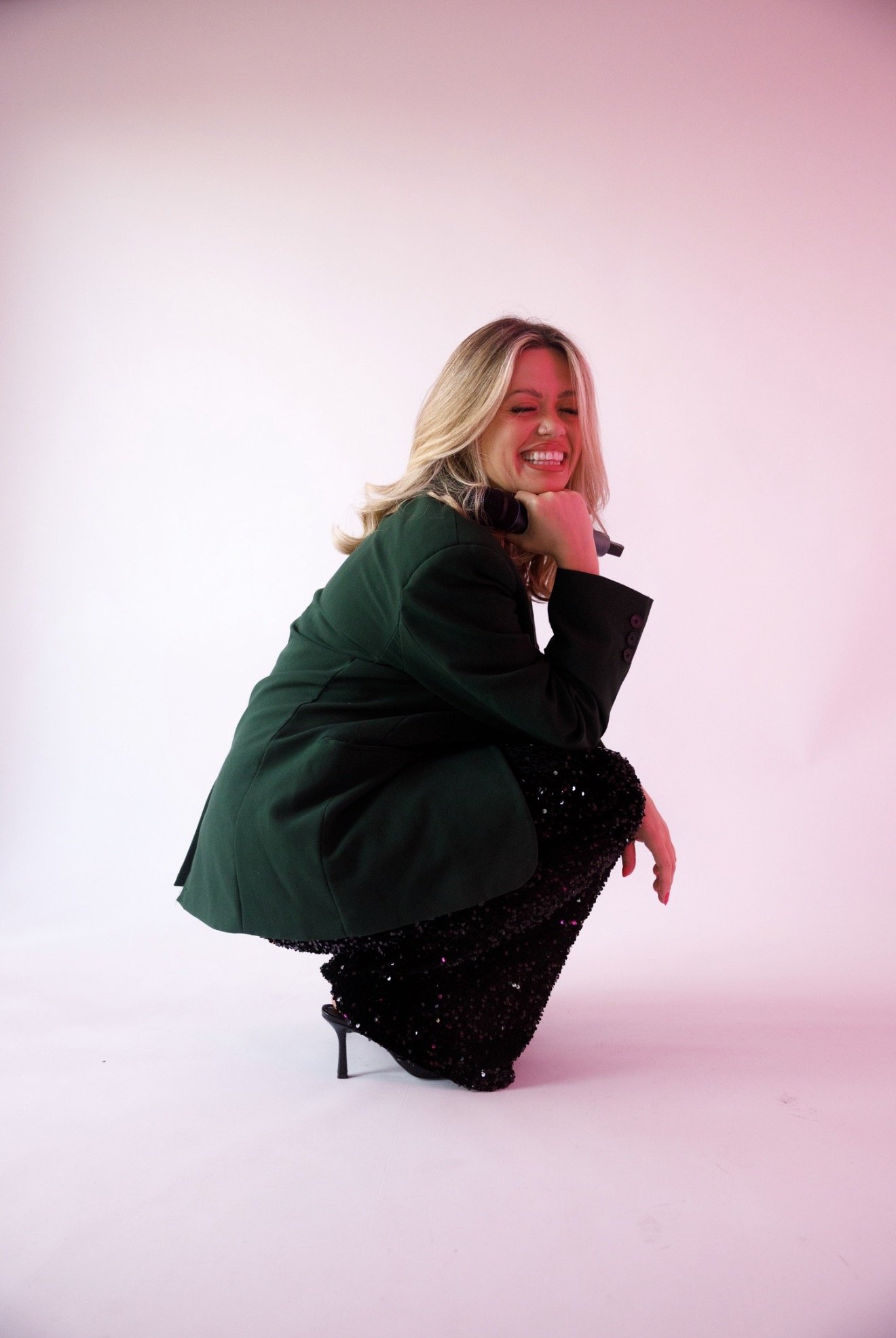 Young woman with blond hair wearing a black sequined dress, black high heels, and a black blazer, sitting on a man's shoulders, laughing with a colorful pink and green background.