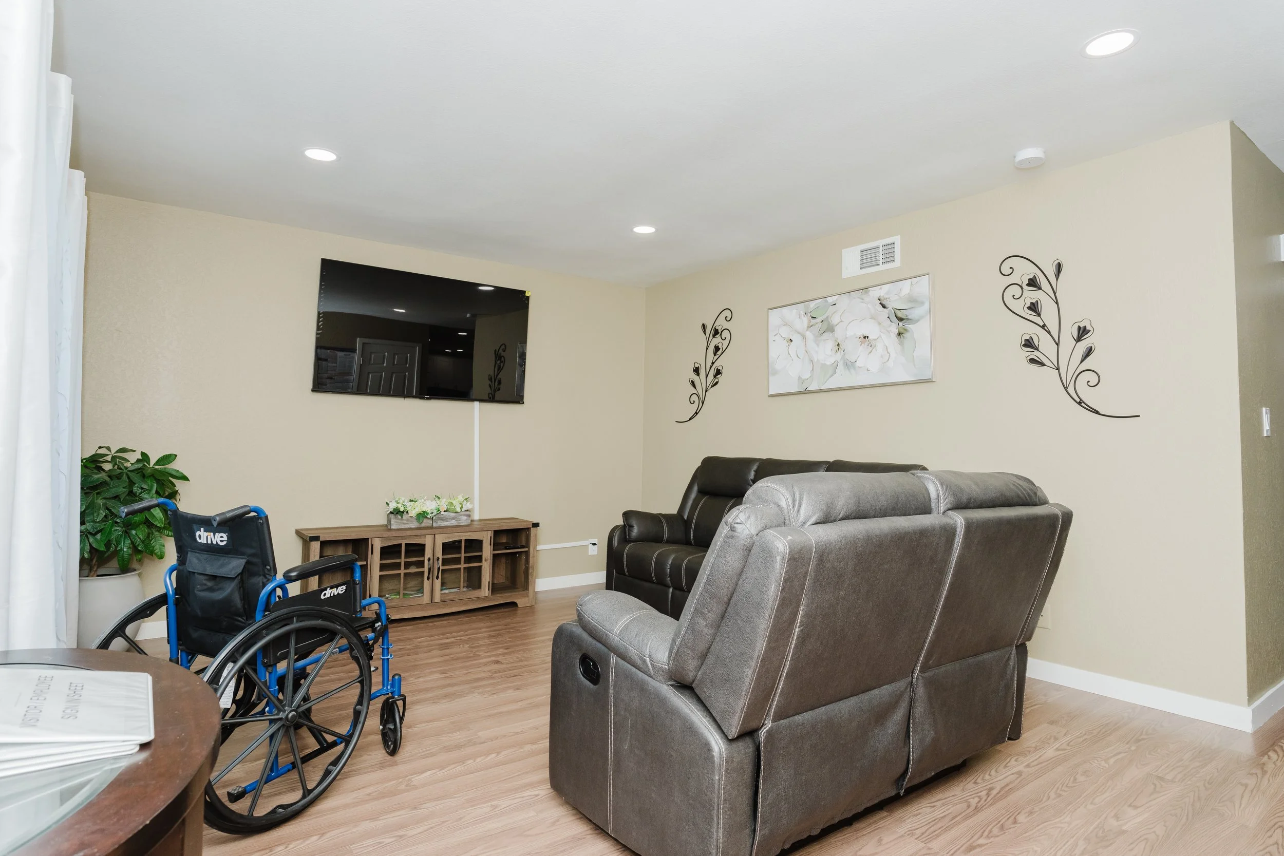 Living room with beige walls, hardwood flooring, a wall-mounted TV, a floral painting, decorative wall art, a large plant, a wheelchair, and a reclining chair.