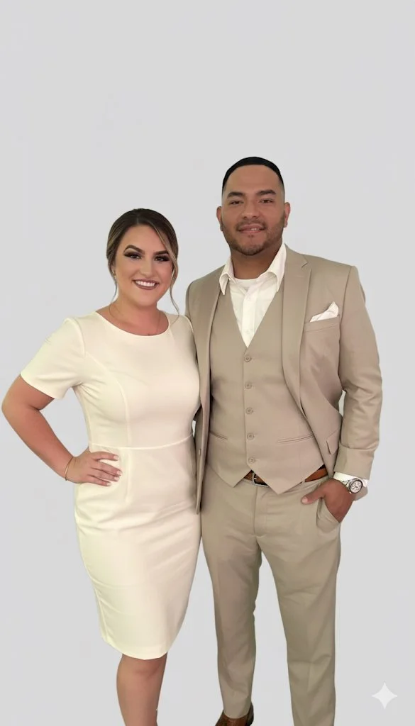 A smiling woman in a white dress standing next to a man in a beige suit against a plain white background.