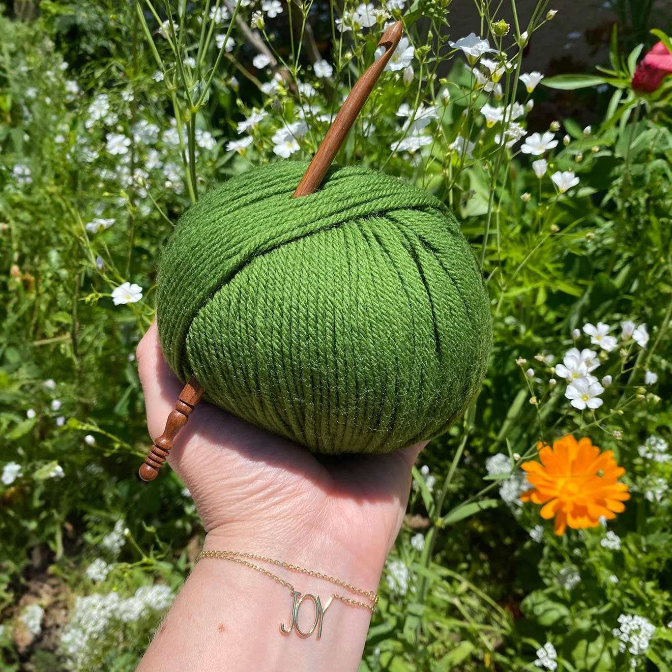 A person's hand holding a ball of green yarn with wooden crochet hook in a garden with white and orange flowers.