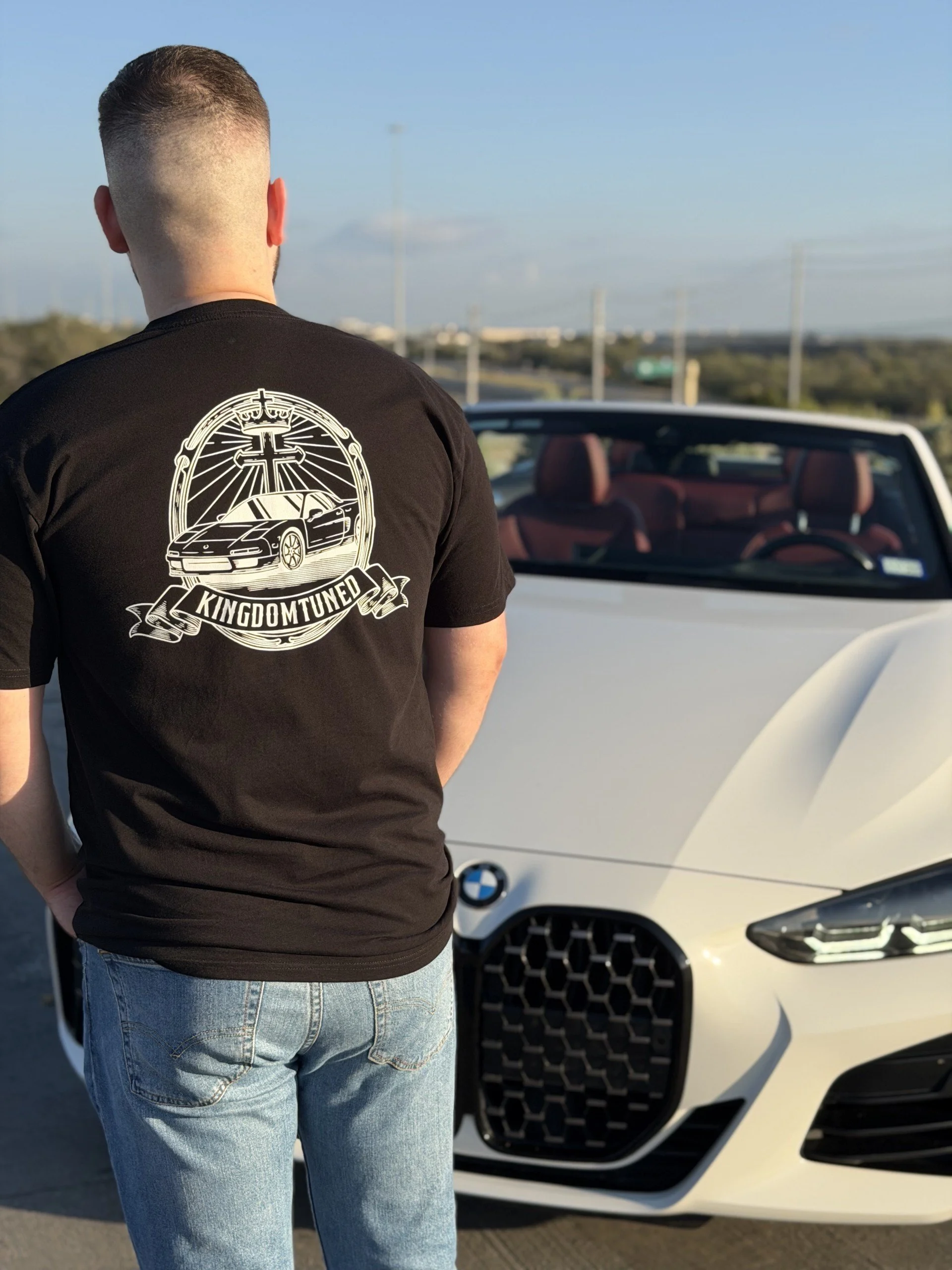 A person with a fade haircut wearing a black t-shirt with a graphic of a car, cross, and the words "Kingdom Tuned" on the back, standing in front of a white BMW convertible with red interior.