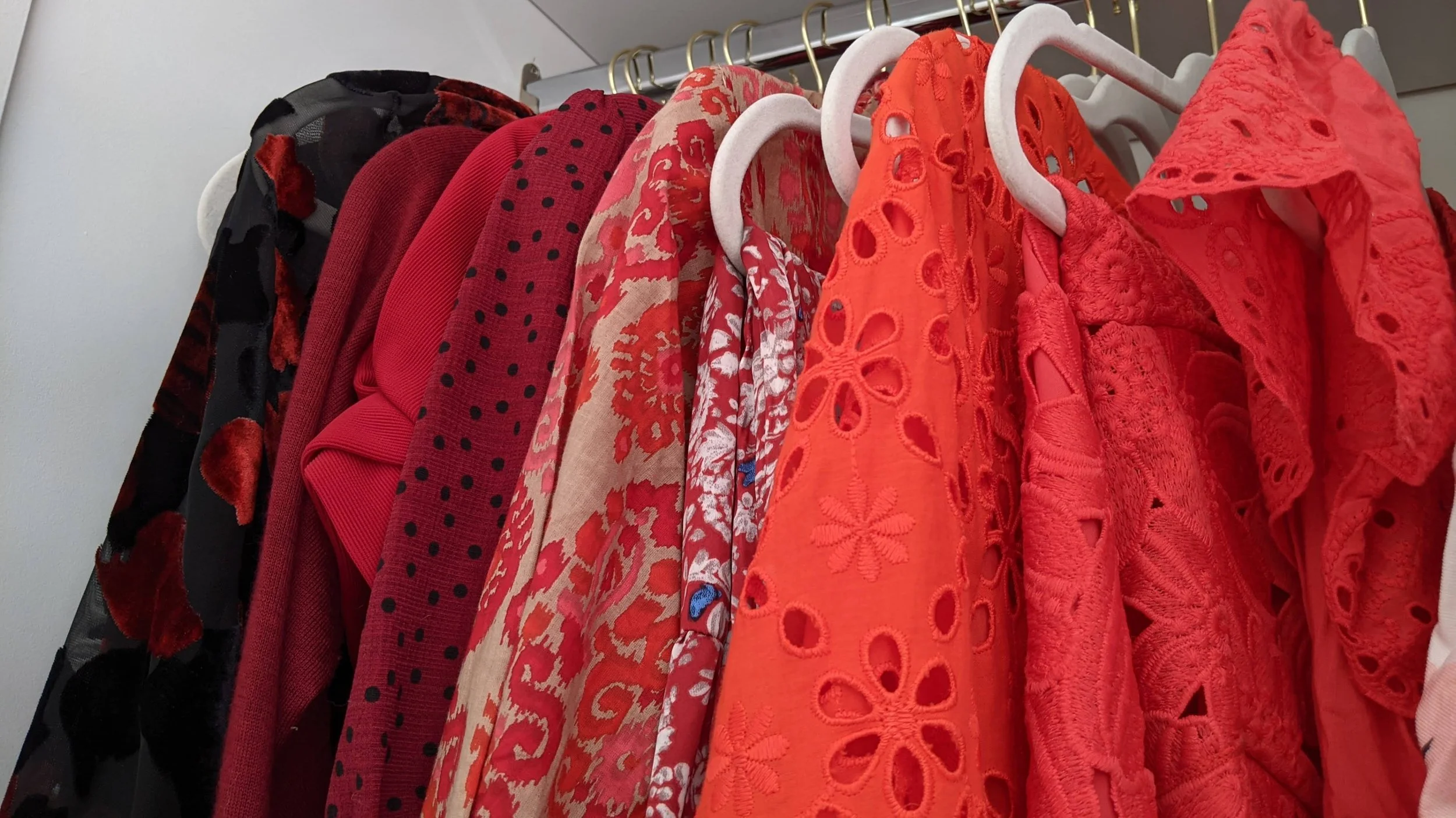 A row of red and black clothing hung on white hangers on a metal rod. The clothes include various patterned fabrics and textures, predominantly in red, with some black accents.