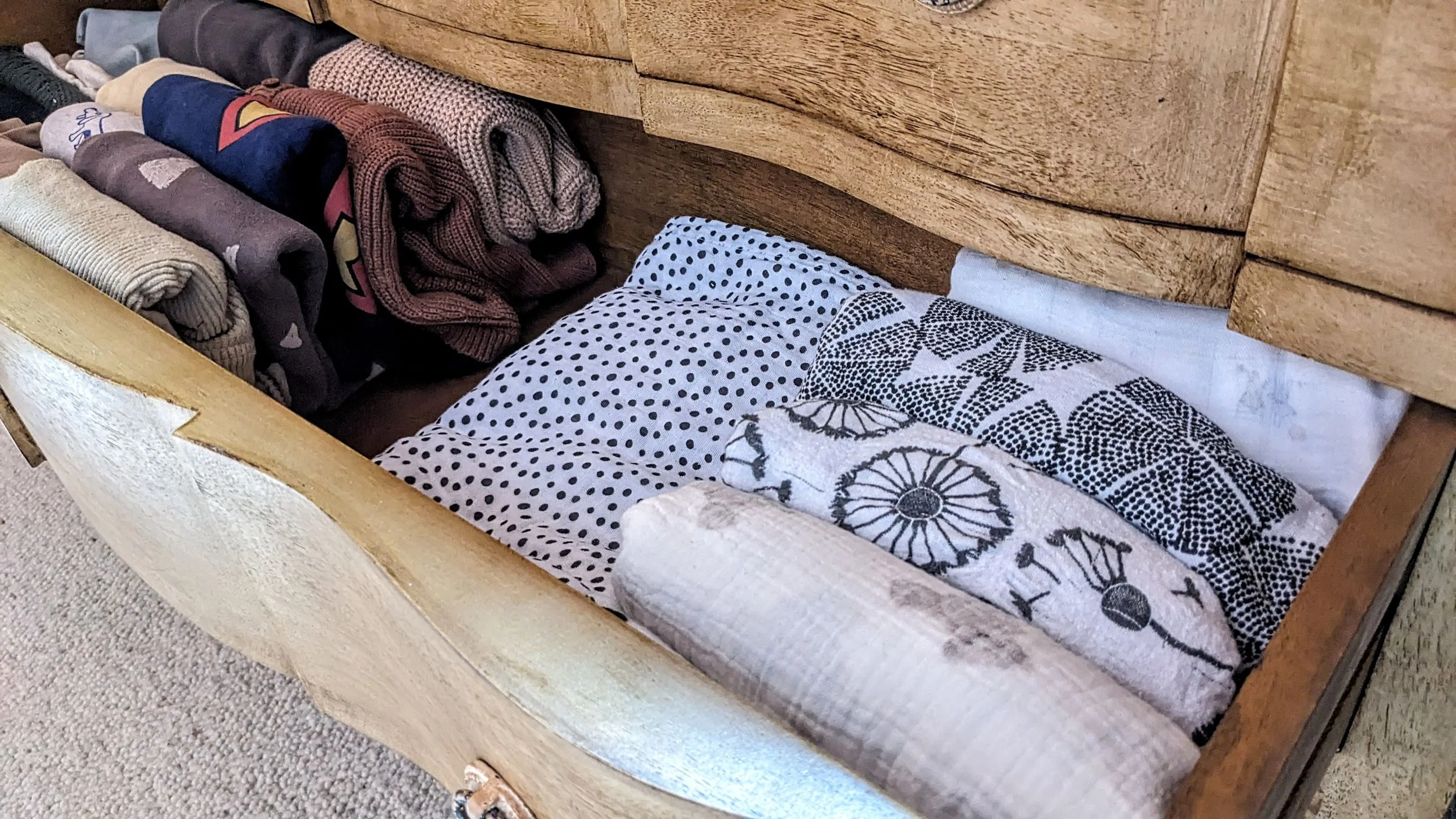 Open wooden drawer with neatly folded clothes and towels inside, including socks, t-shirts, and patterned linens.