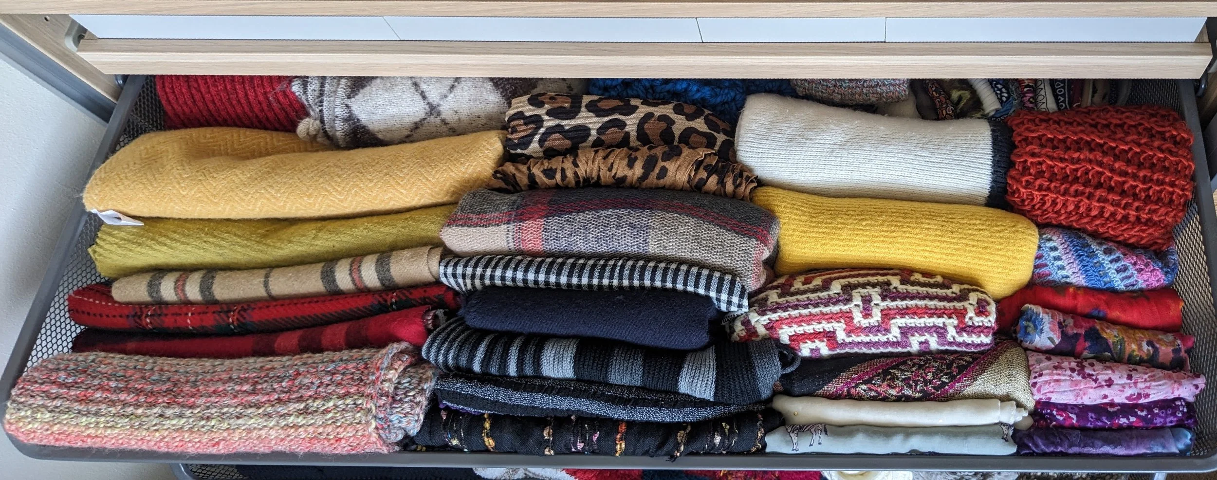 A mesh metal drawer filled with neatly folded scarves and sweaters in various colors and patterns, including plaid, floral, and knitted designs.