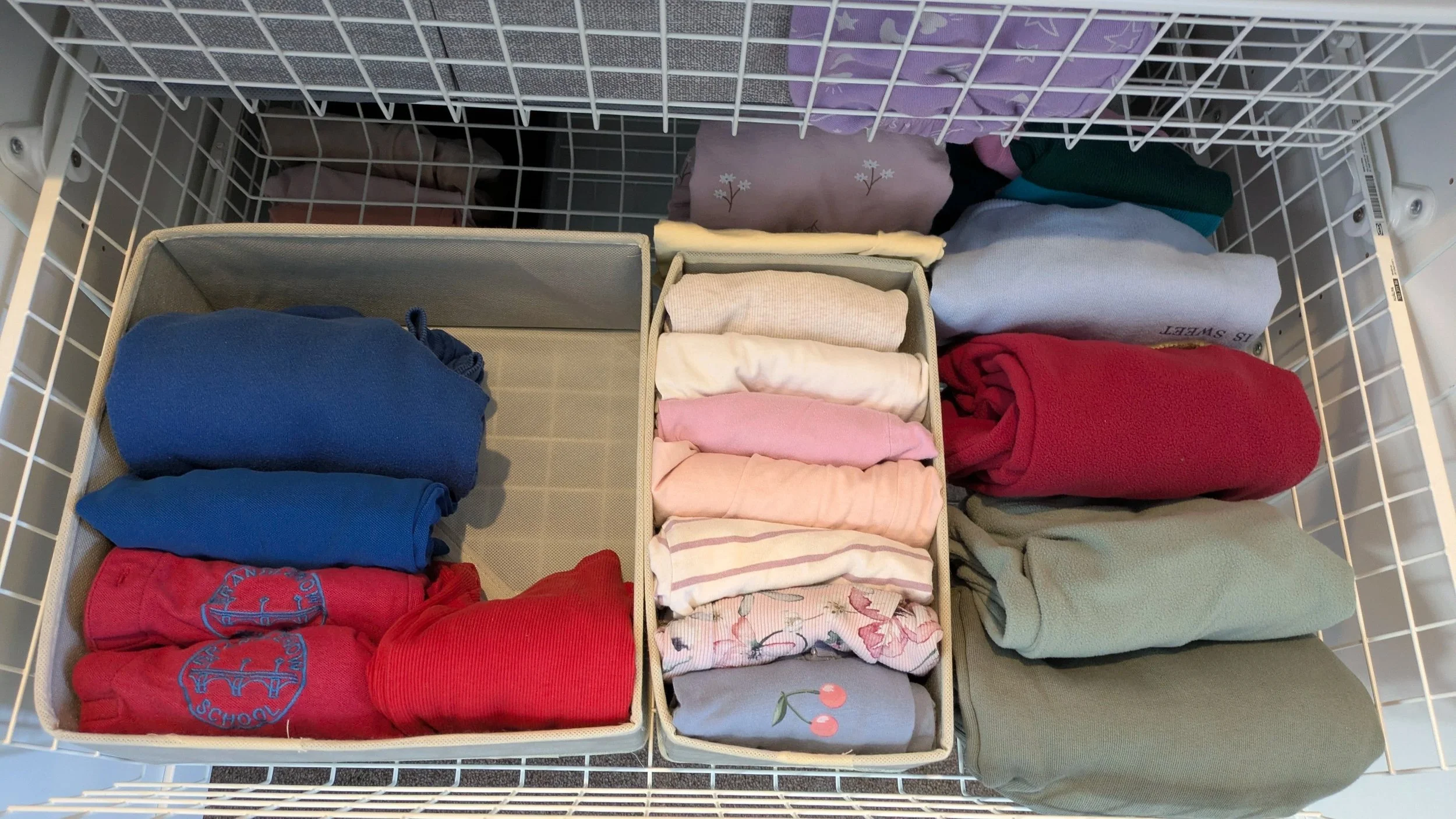 Organized drawer with folded clothes, including t-shirts, socks, and pants, in separate beige and fabric storage boxes, with additional clothes hanging or folded nearby.