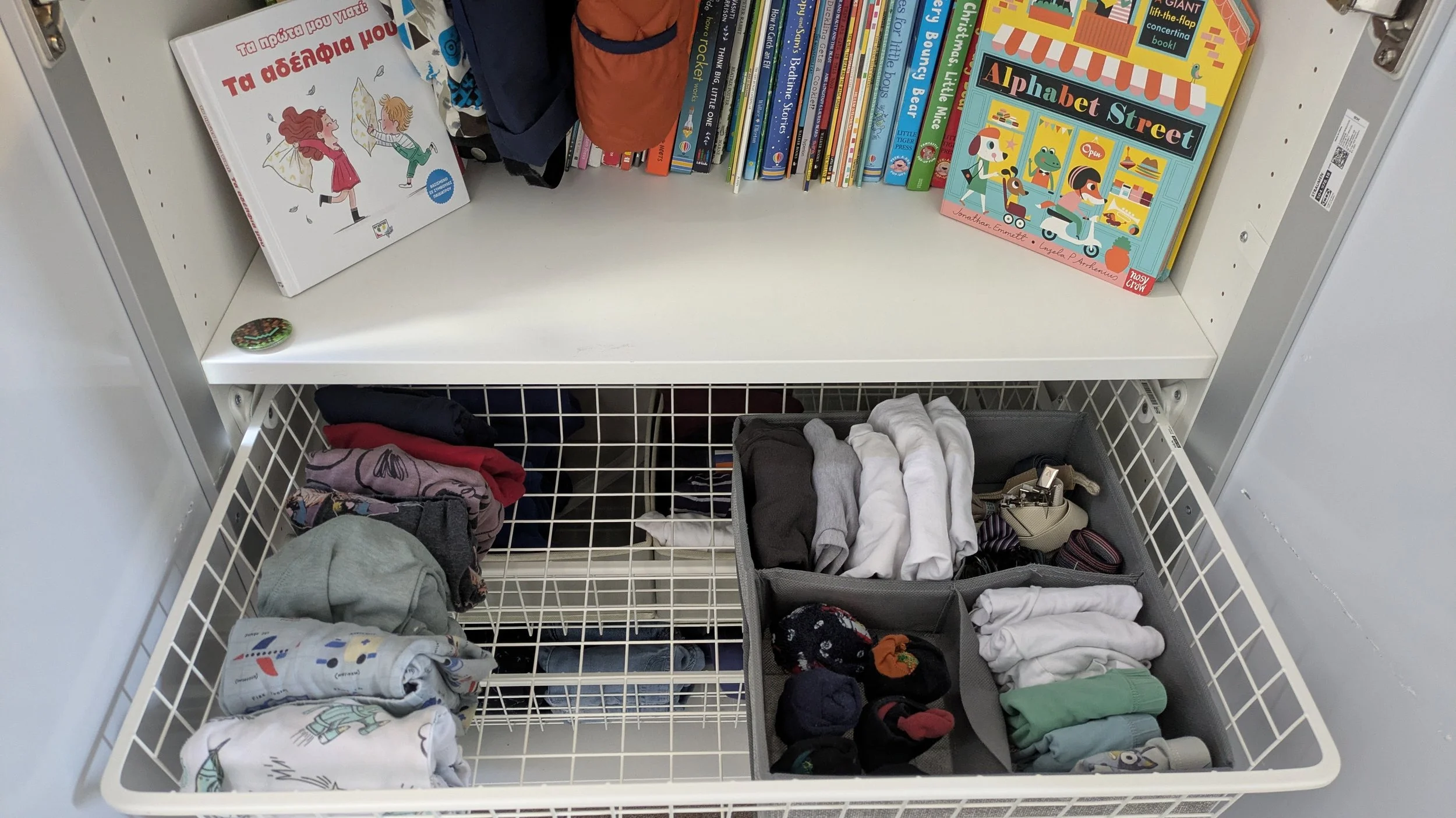 View of a white wire drawer filled with folded clothes and socks, with a white shelf above holding children's books about dragons, John Lewis, and alphabet street, with backpacks hanging behind.
