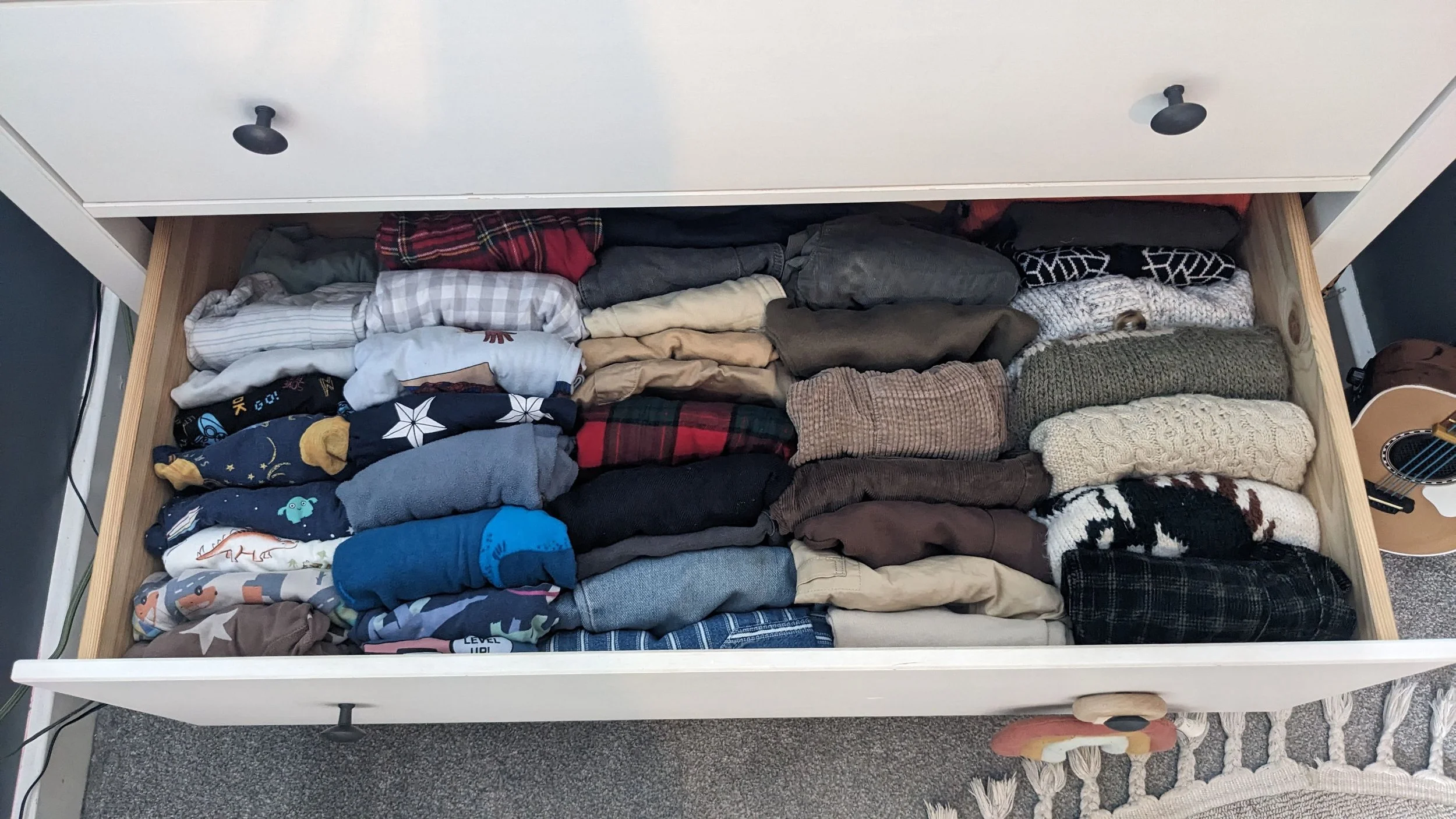 Open drawer containing neatly folded clothes, including sweaters, shirts, and pants, with part of a guitar visible on the right side and a patterned rug at the bottom.