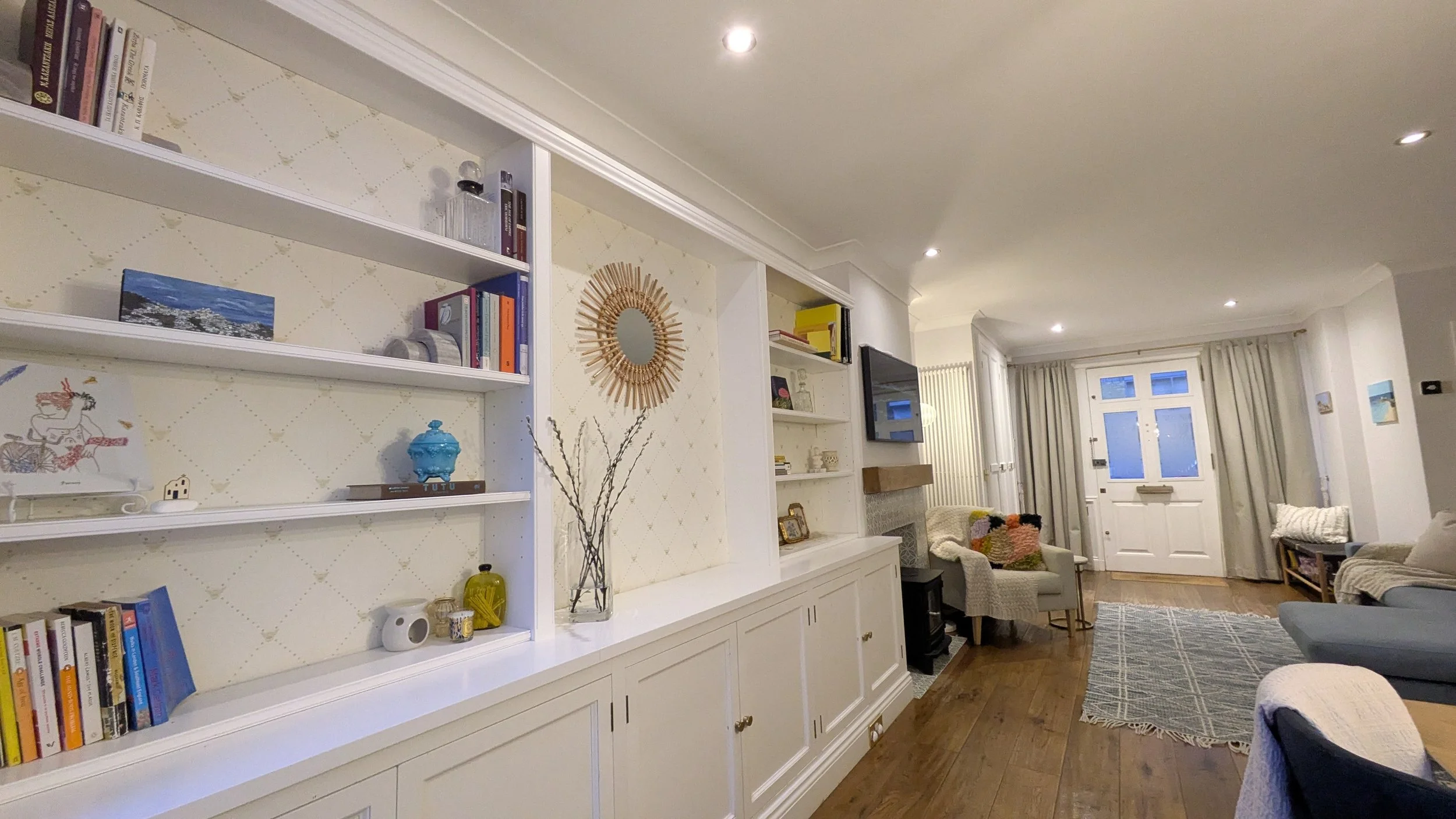 Living room with white built-in shelves filled with books and decorative items, cream armchair, patterned rug, large window with curtains, and a white front door with small window panels.