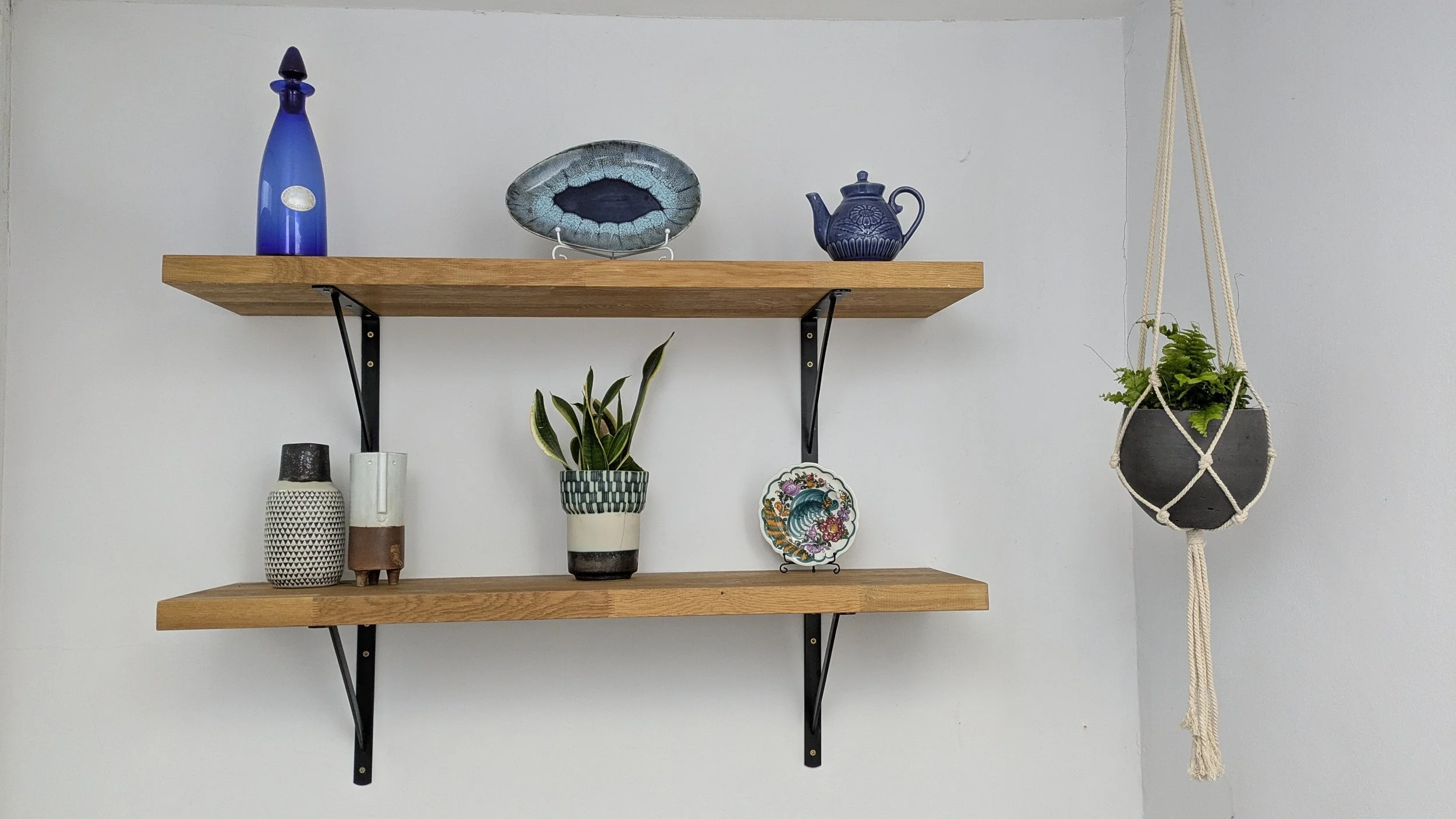Two wooden shelves with black metal brackets hold decorative vases, plates, and a teapot. A hanging planter with green plants is on the right side of the shelves.