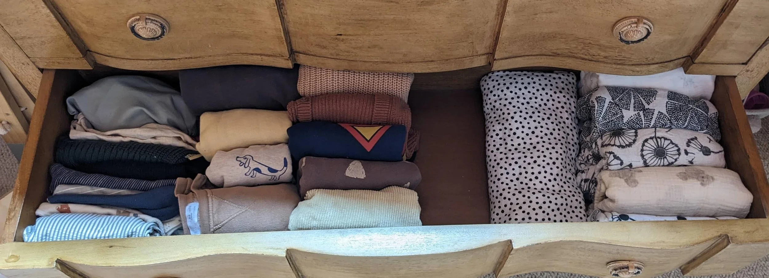 Open wooden drawer filled with folded clothes, including shirts, sweaters, and linens of various colors and patterns.