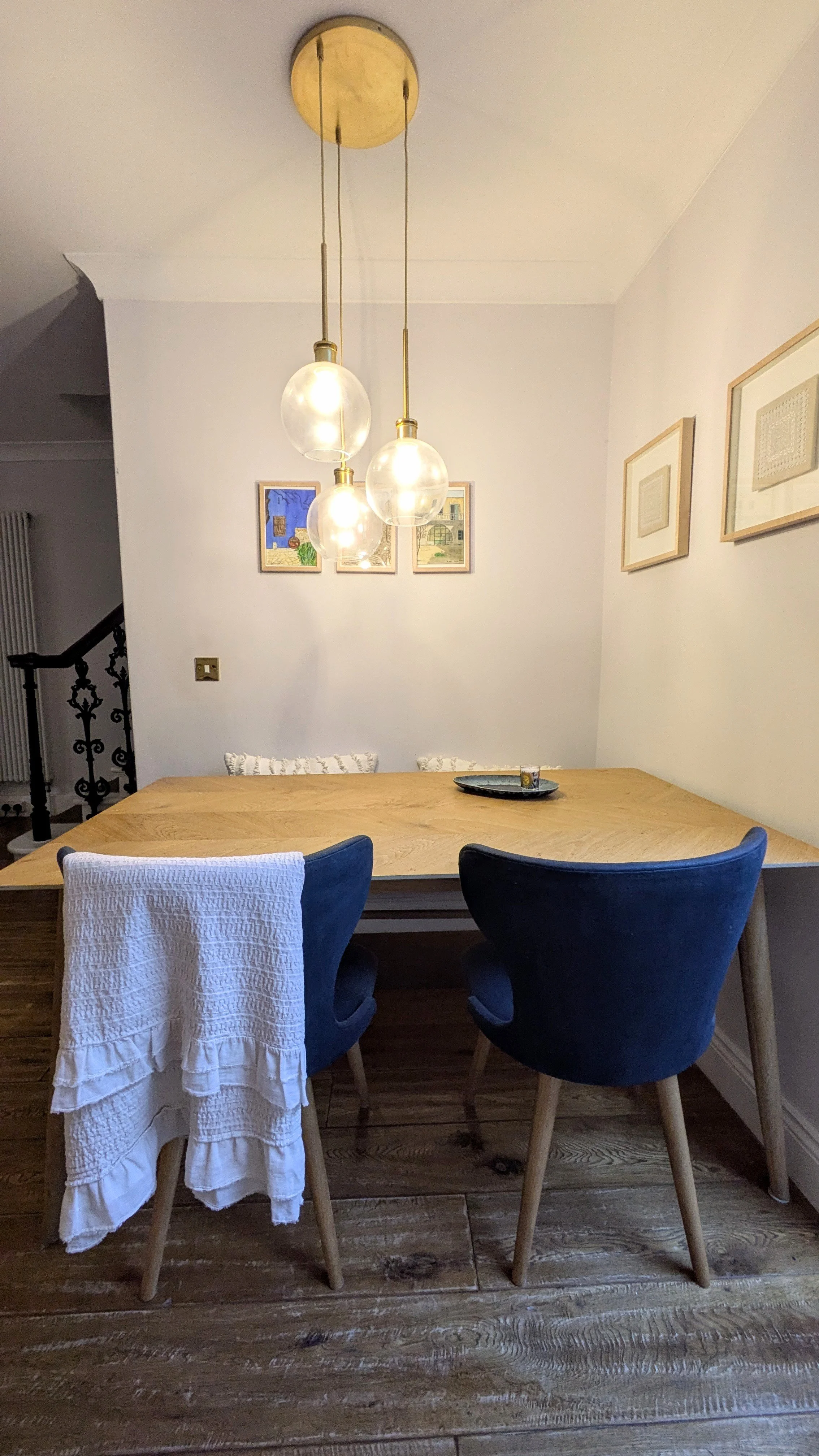 A dining area with a wooden table, two navy blue chairs, and a white textured cloth draped over one chair. There are framed pictures on the wall, a modern hanging light fixture with three glass globes above the table, and a wooden floor.