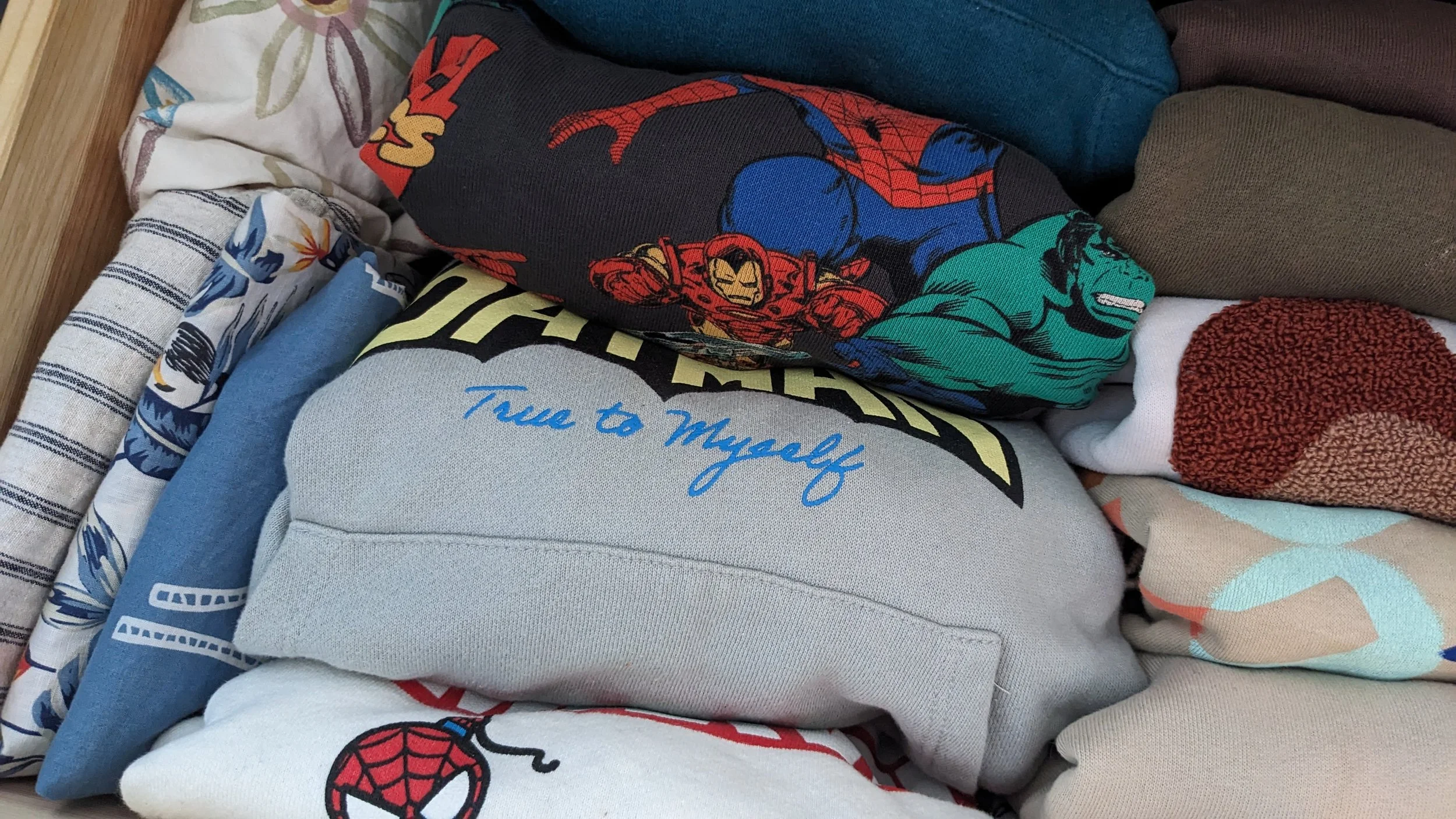 A collection of folded clothing items featuring various superhero and cartoon designs, including Spider-Man, Iron Man, Hulk, and others.