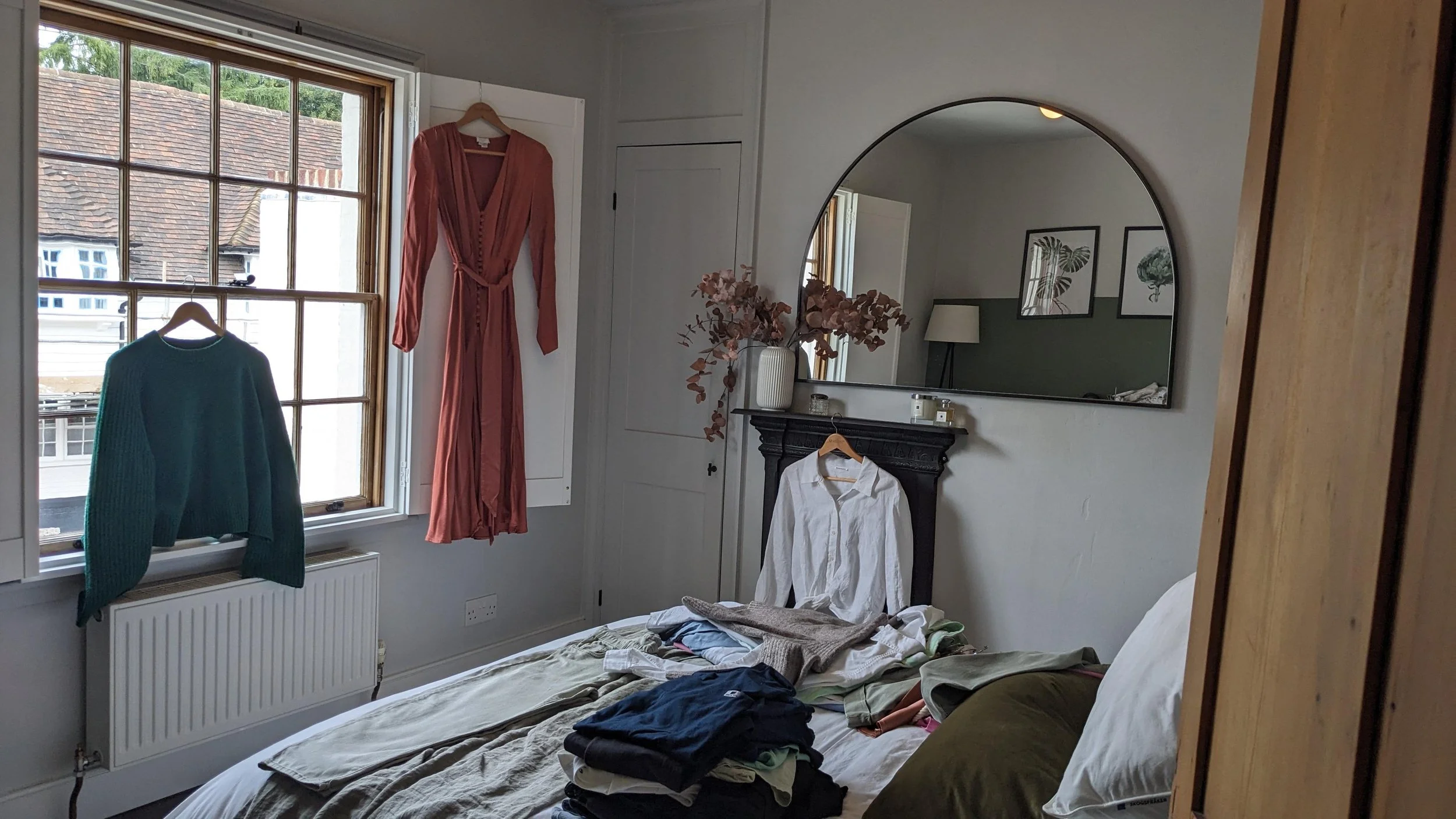 A bedroom with an unmade bed, two hanging clothes (a pink dress and a teal sweater), a large mirror above a black fireplace, and a window showing rooftops outside.