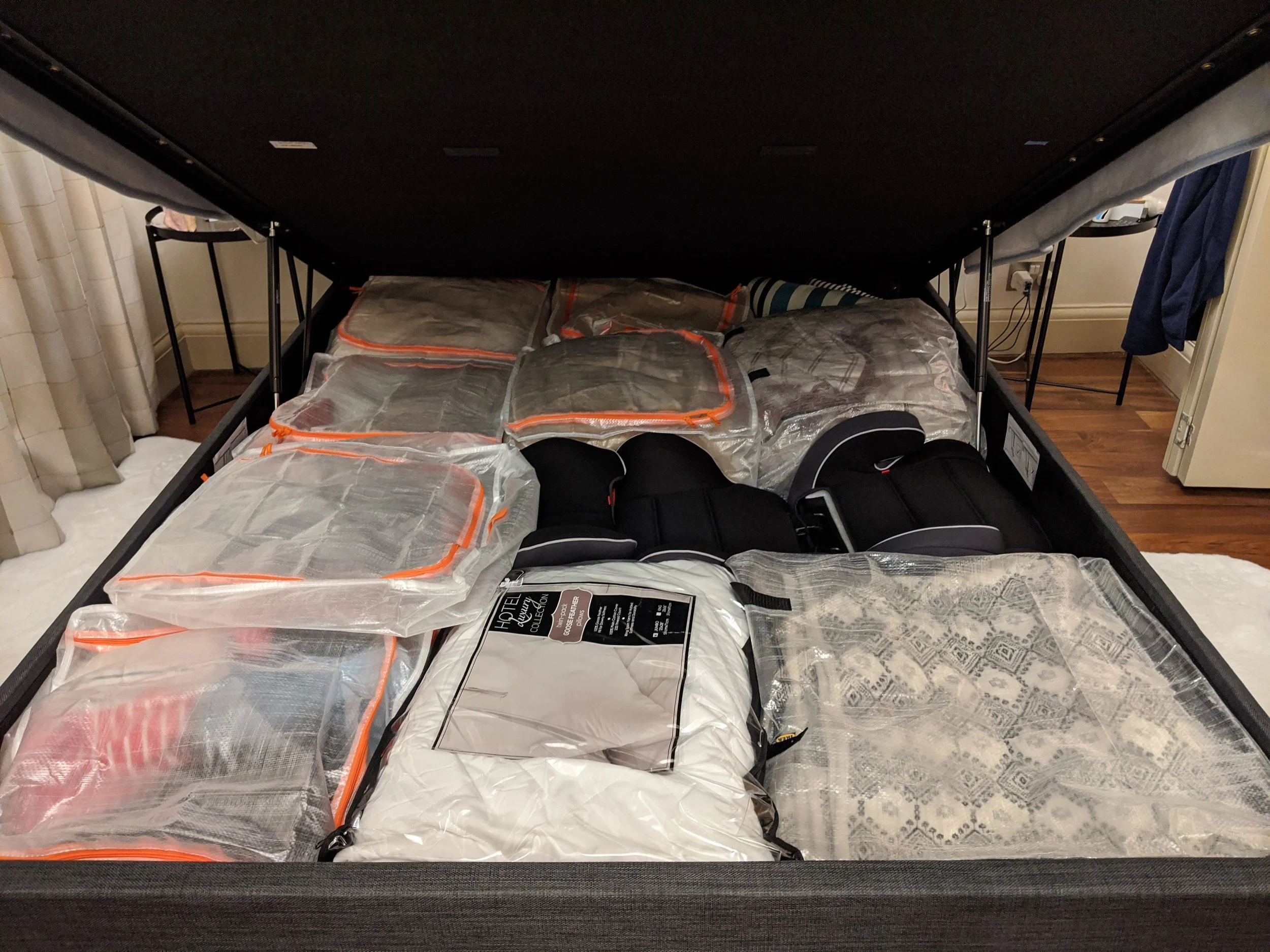 A bed with a black frame containing various packaged items, including comforters, pillows, and possibly clothing, inside transparent storage bags with orange zippers.