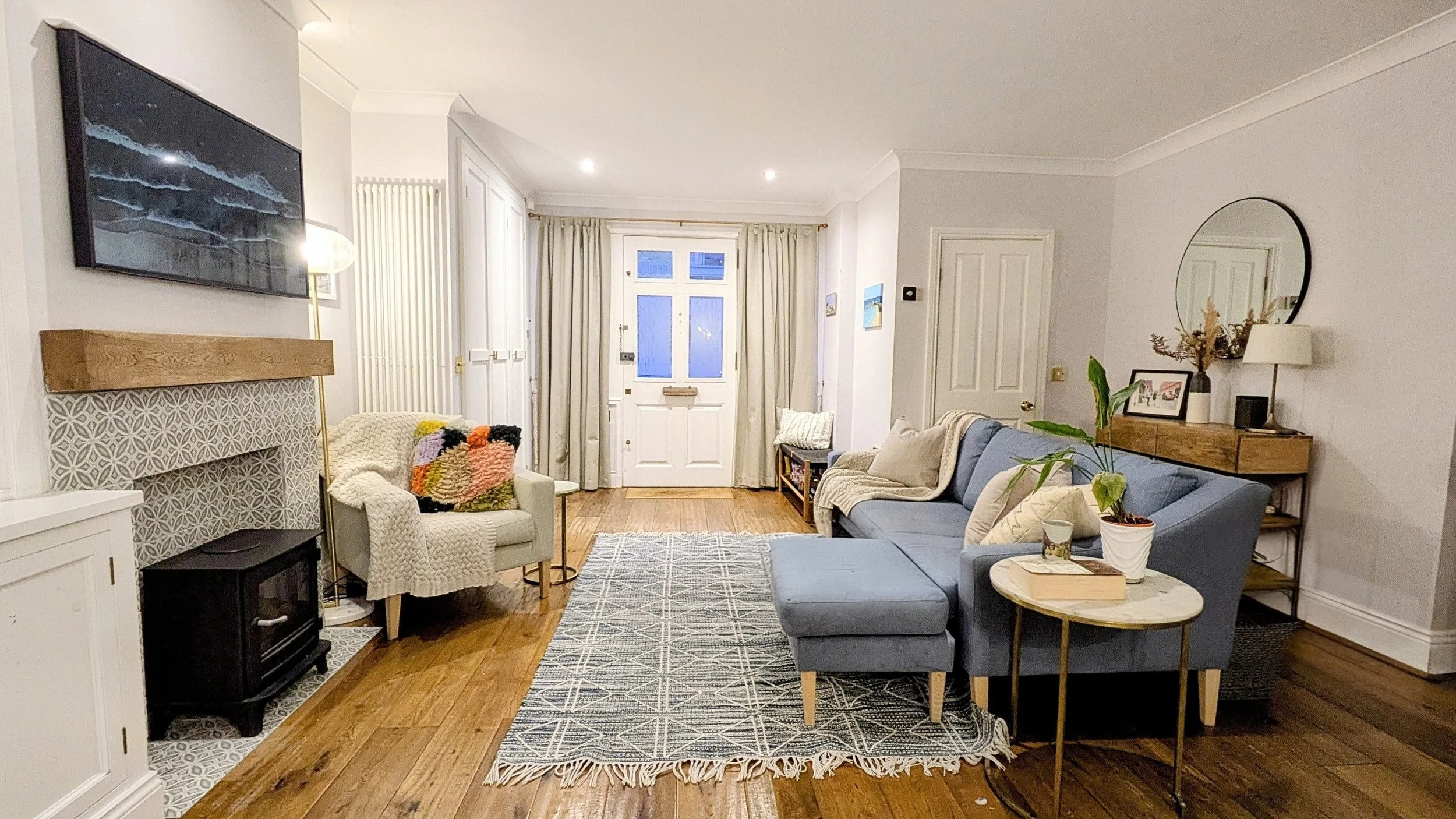 Living room with a gray sofa, a blue armchair, a patterned rug, a white fireplace, a wall-mounted TV, and a wooden side table with plants and framed photos, with curtains and a door in the background.