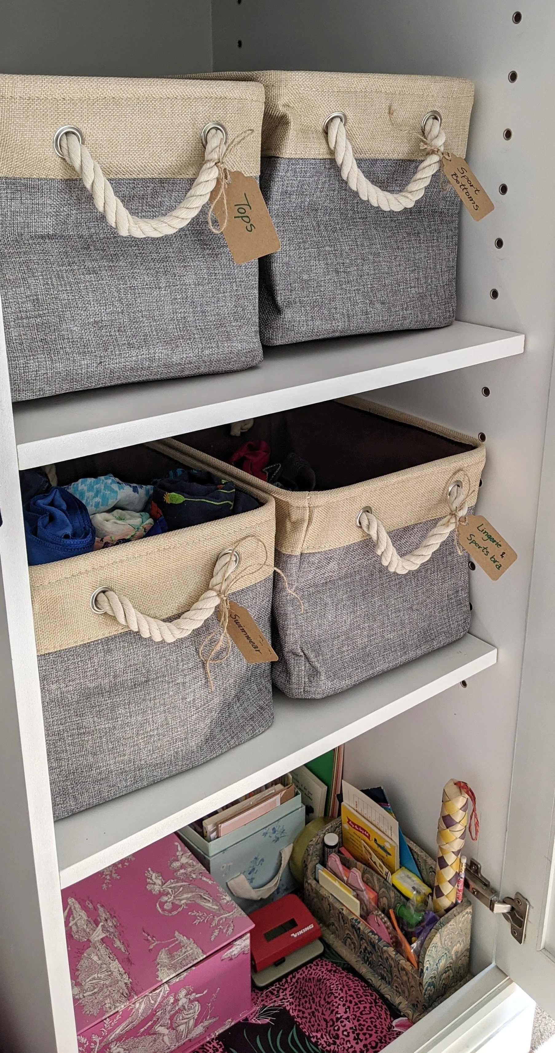 A closet with shelves containing fabric storage bins labeled for different items such as tops, lingerie, and sports bras. The bottom shelf has various boxes, books, and miscellaneous items.