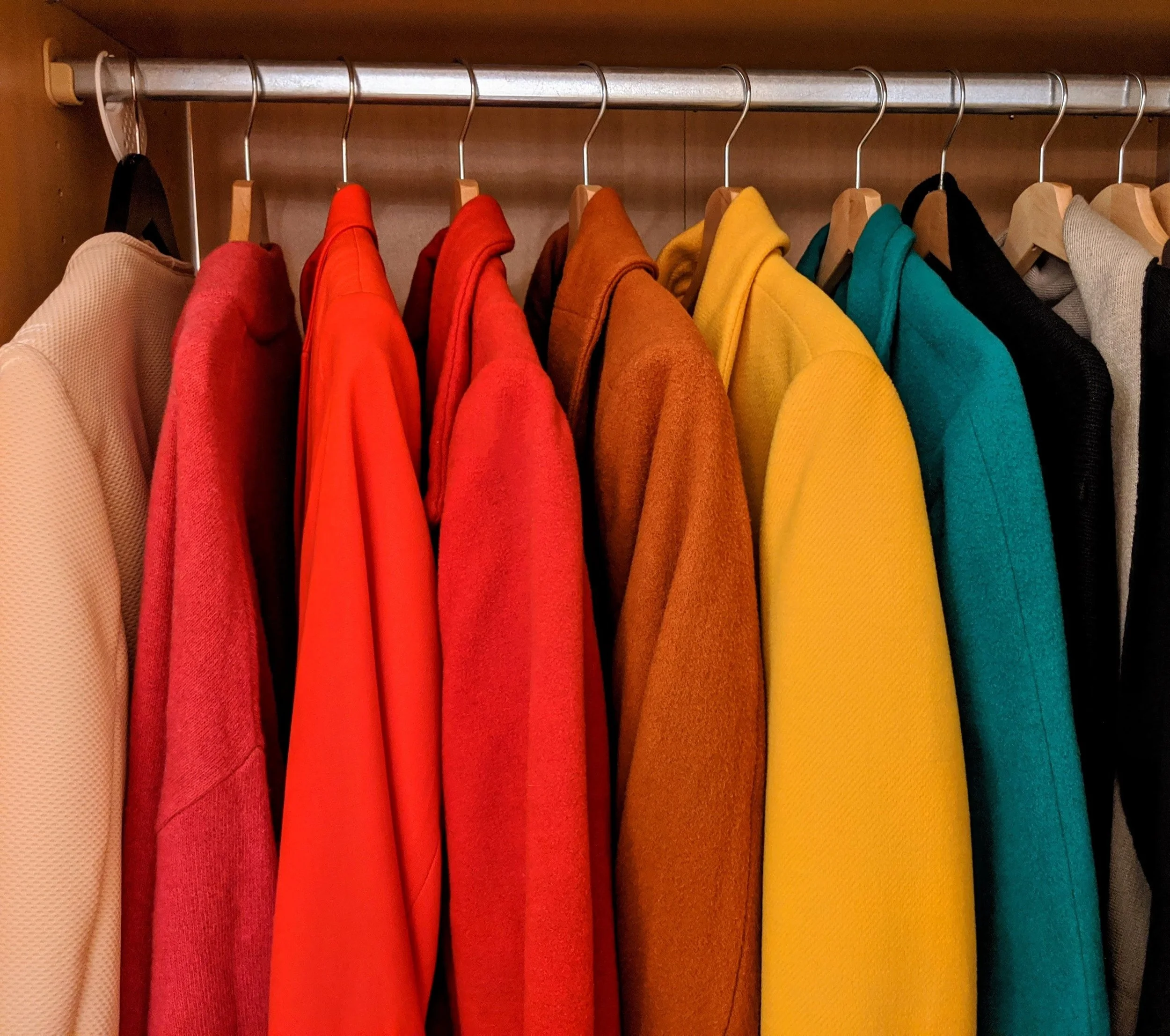 A closet with a variety of colorful shirts hanging on a silver rod: beige, red, orange, brown, yellow, teal, black, and gray.