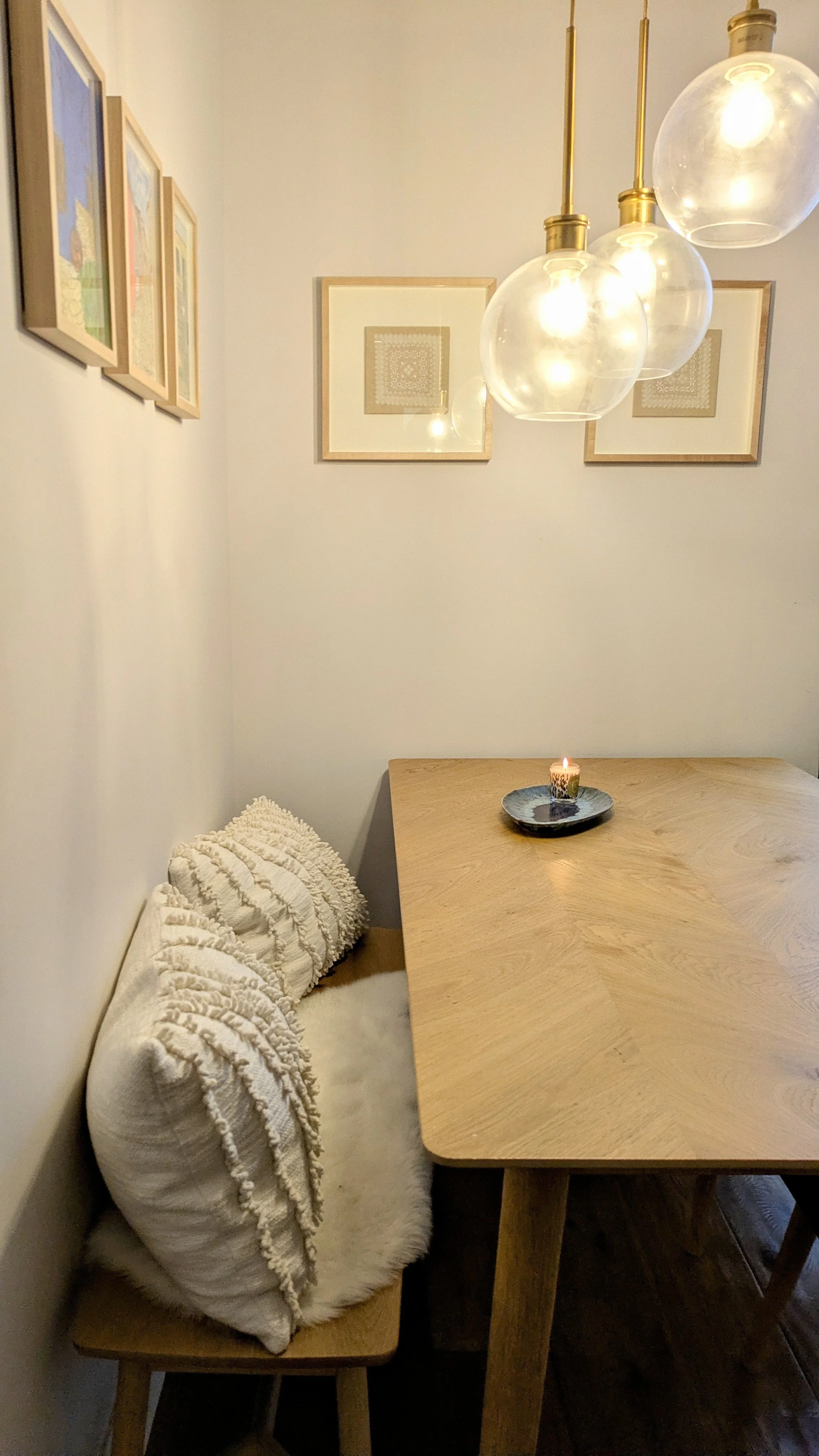 A cozy dining nook with a wooden table, a bench with textured white throw pillows, a small candle on a black plate, framed artwork on white walls, and hanging globe pendant lights.