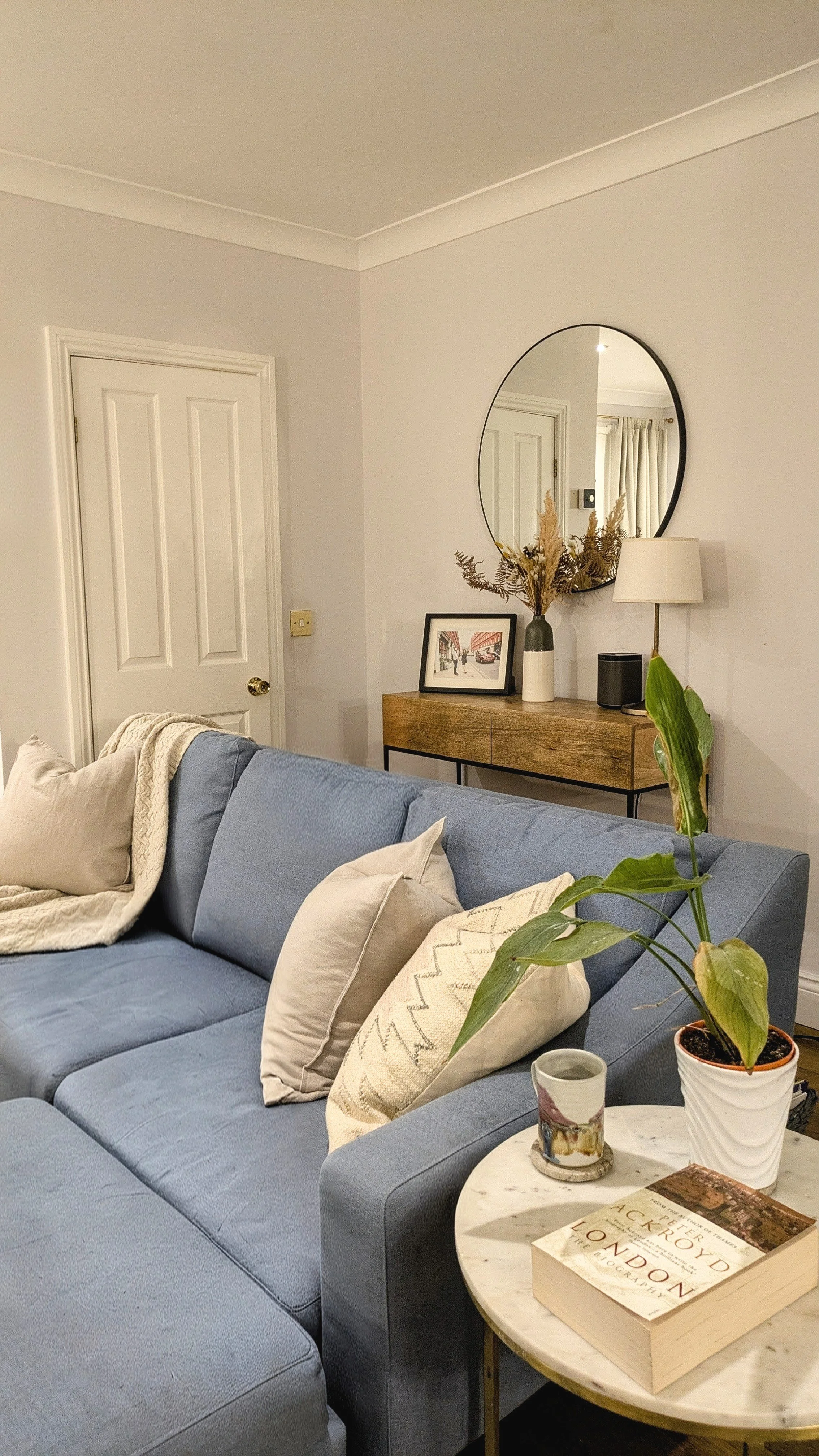 A cozy living room with a blue sofa decorated with beige and patterned throw pillows, a round marble side table with plants, a book, and a mug, a wooden console table with a framed picture, a vase with dried flowers, a lamp, and a mirror hanging abov