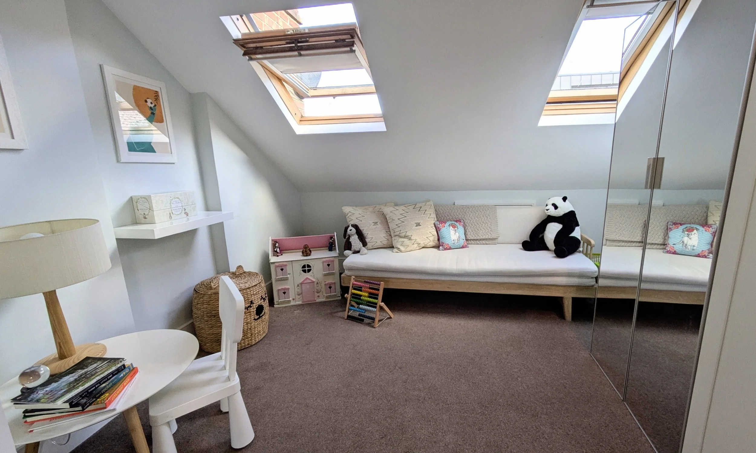 A cozy attic room with skylight windows, a white sofa with pillows and stuffed animals, a small dollhouse, a wicker basket, a small table with books, and a mirrored closet door.