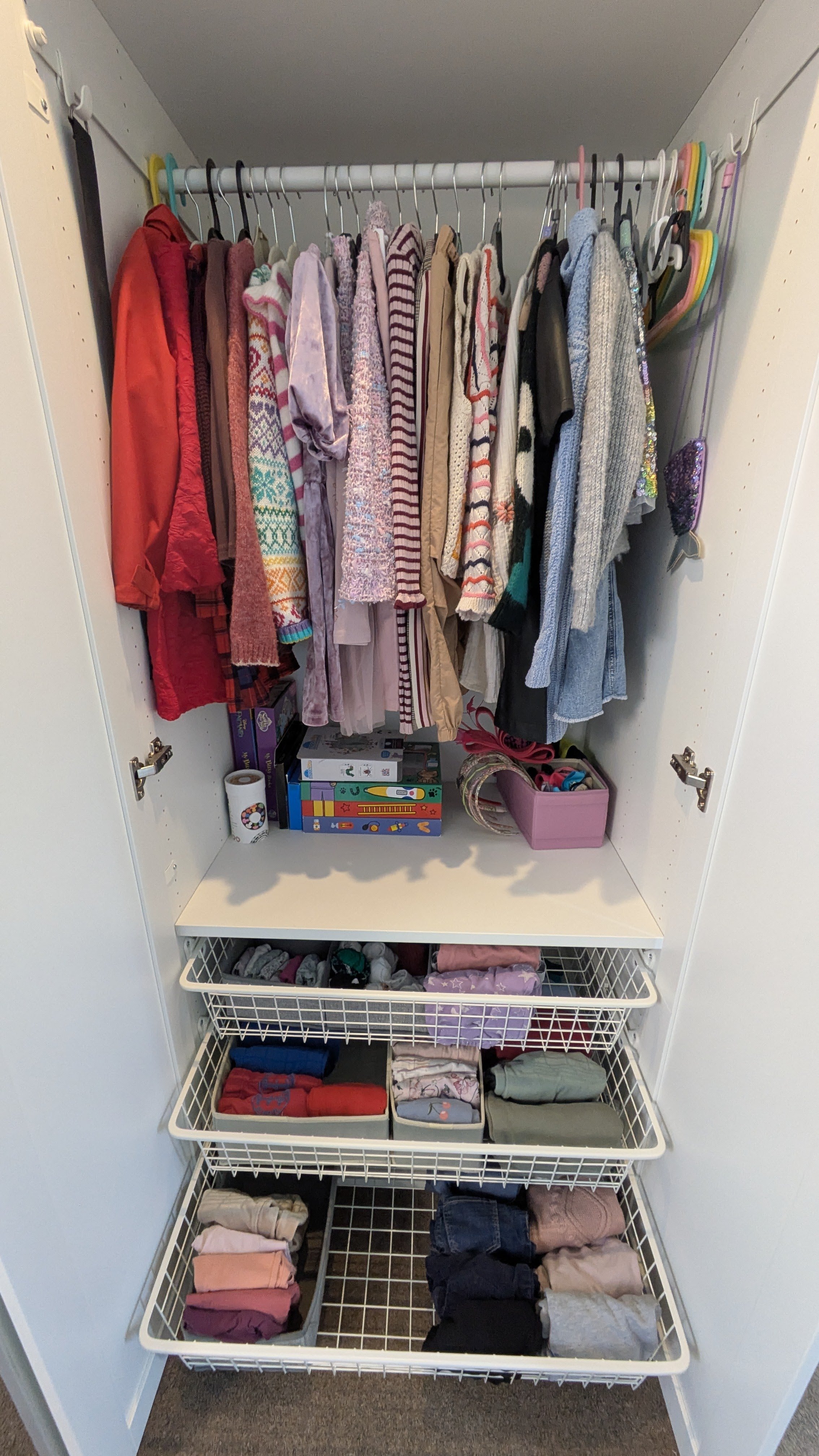 Inside a closet, hanging on a white rod are children's clothes including jackets, sweaters, and shirts in various colors and patterns. Below are three open shelves filled with folded clothing items and bins holding socks and other small accessories.