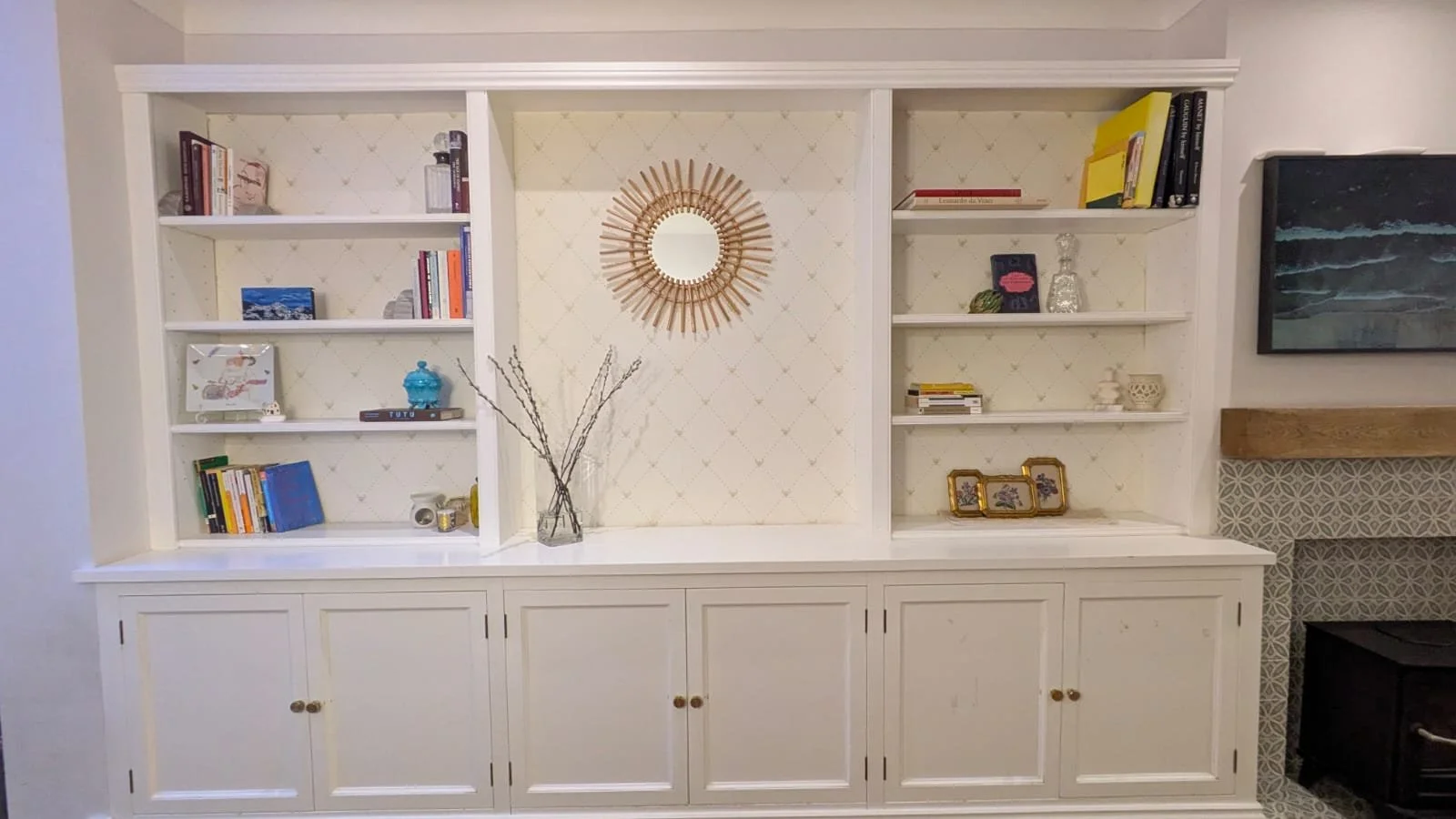 A white built-in bookshelf with decorative objects, framed photos, books, and a vase with branches, and a mirror on the wall behind the shelf.