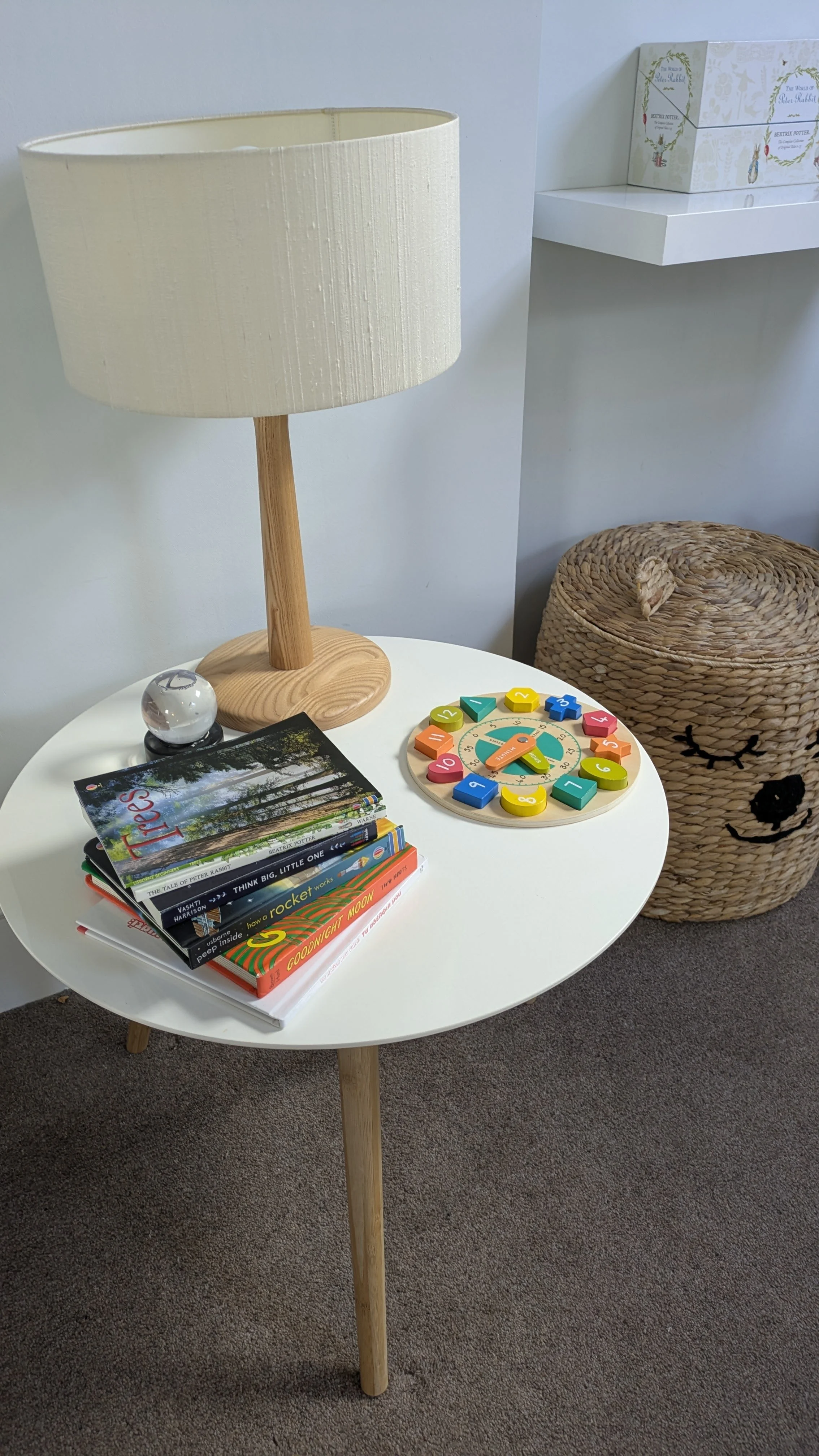 White side table with wooden legs, holding a lamp with a beige shade, a globe, a stack of children's books, and a colorful educational clock toy. In the background, there is a woven basket with a lid and a white shelf with a decorative box.
