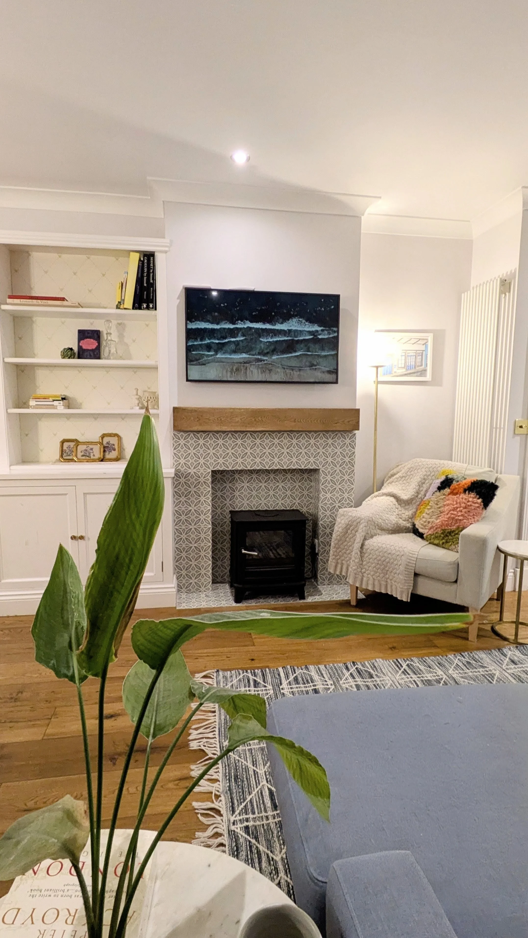 Living room with a white armchair, a blanket, colorful pillows, a fireplace with patterned tiles, a mounted TV above the fireplace, a lamp, built-in shelves with books and decorations, a floor plant, a rug, and wooden floors.