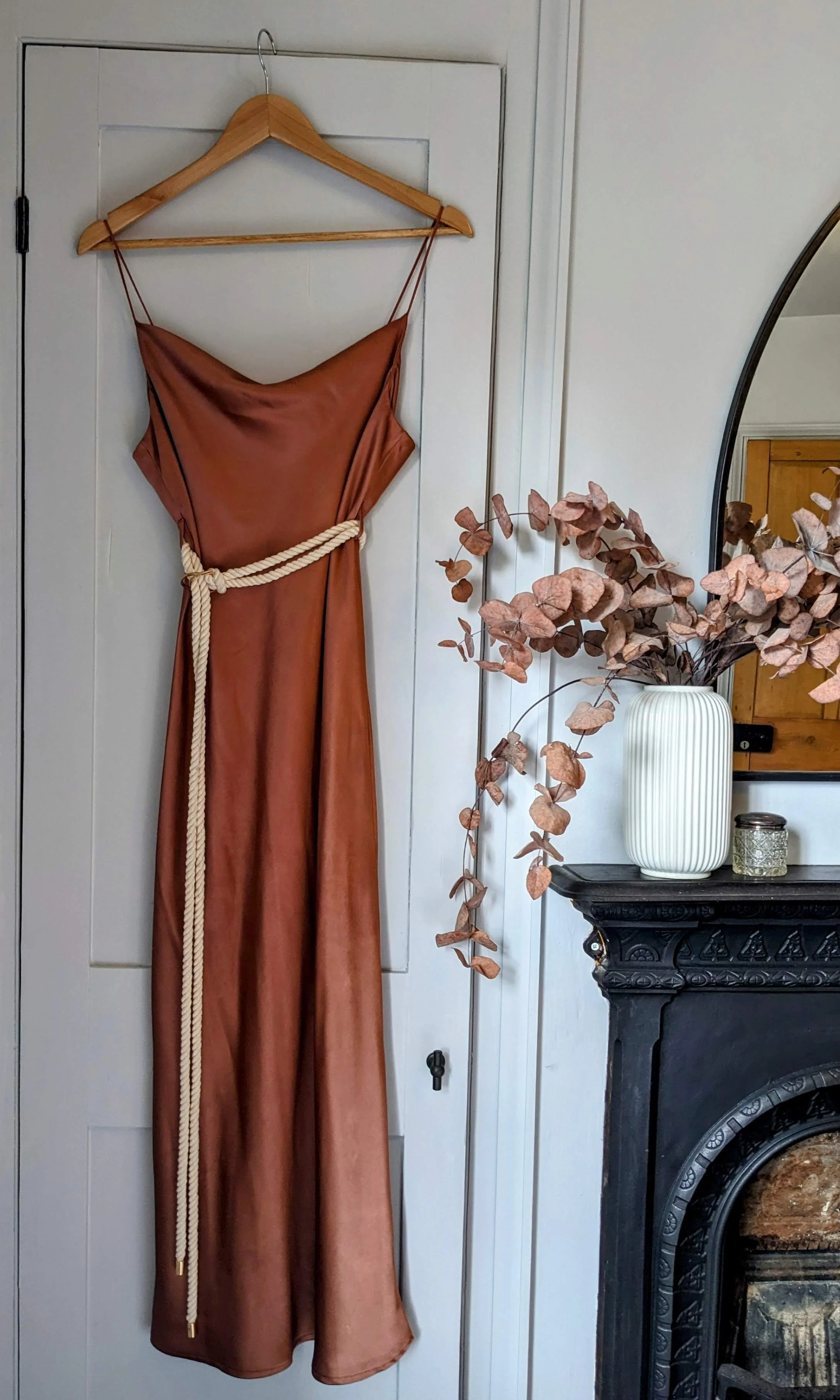 A rust-colored satin dress hangs on a wooden hanger on a white door, with a white rope tied around the waist. Next to it is a white vase with dried pinkish-brown leaves on a black mantlepiece, with part of a mirror and a wooden piece of furniture in