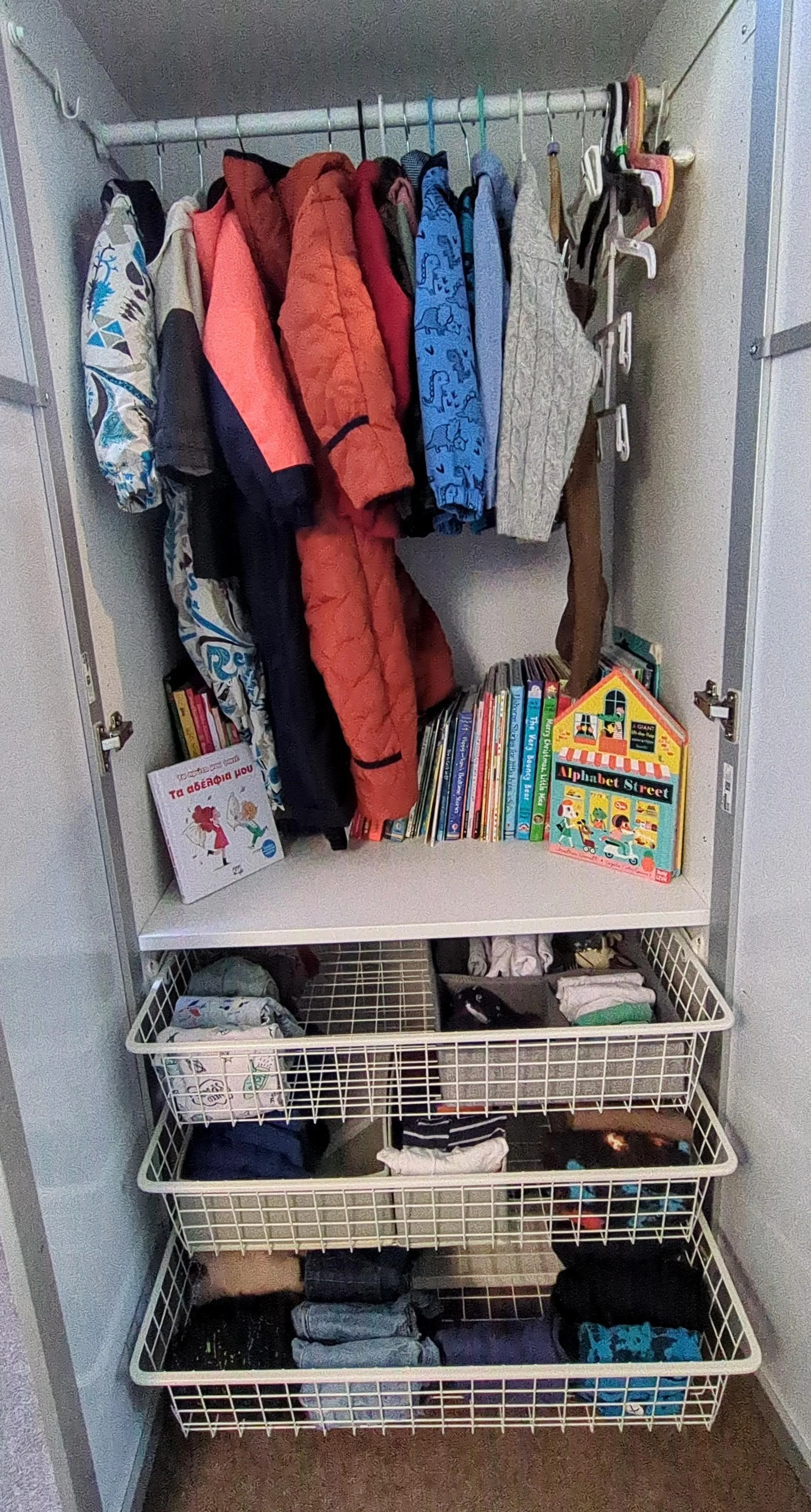 Open closet with hanging jackets and sweaters, shelves with children's books, and wire baskets containing folded clothing and toys.
