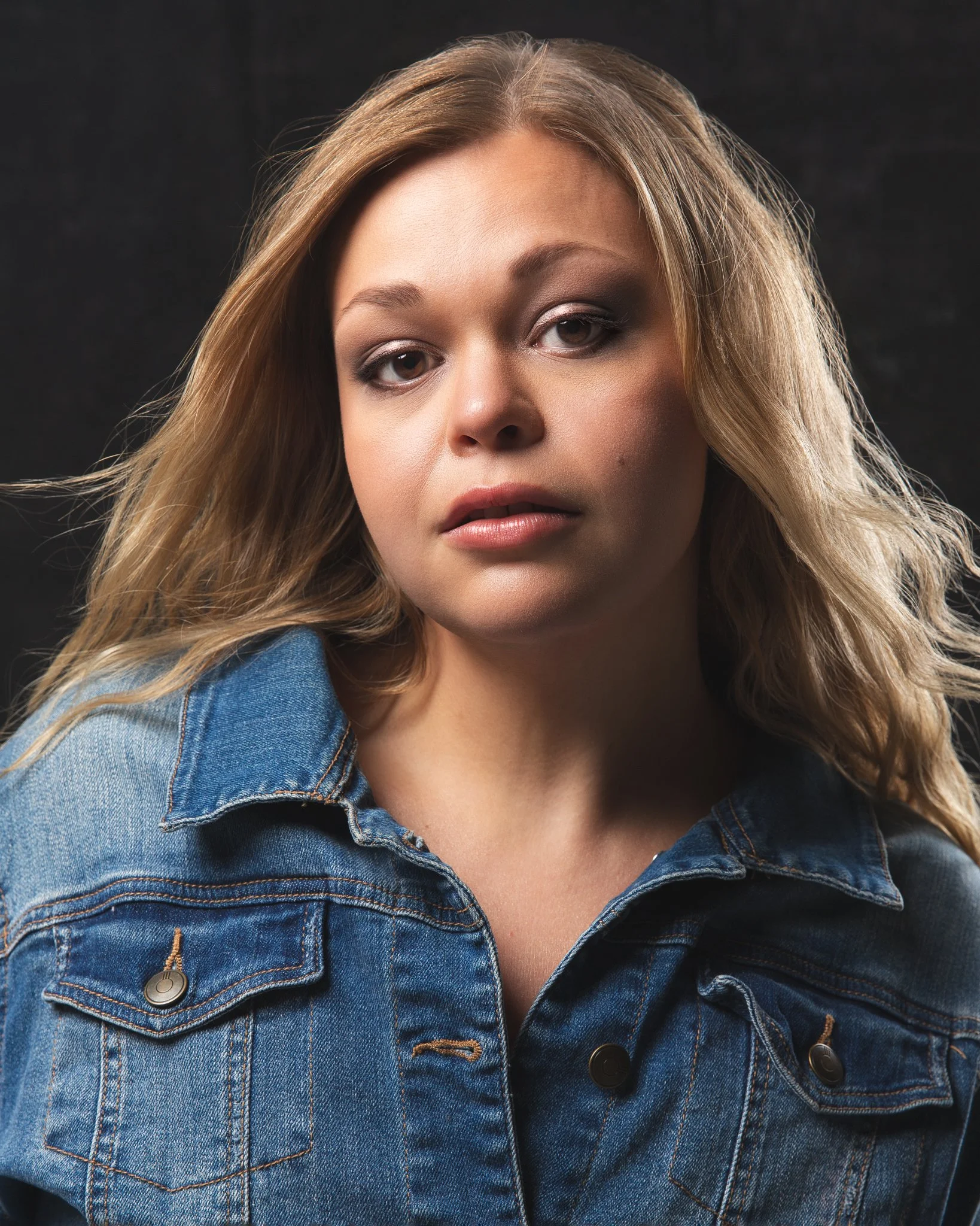 A woman with blonde hair wearing a denim jacket against a dark background.
