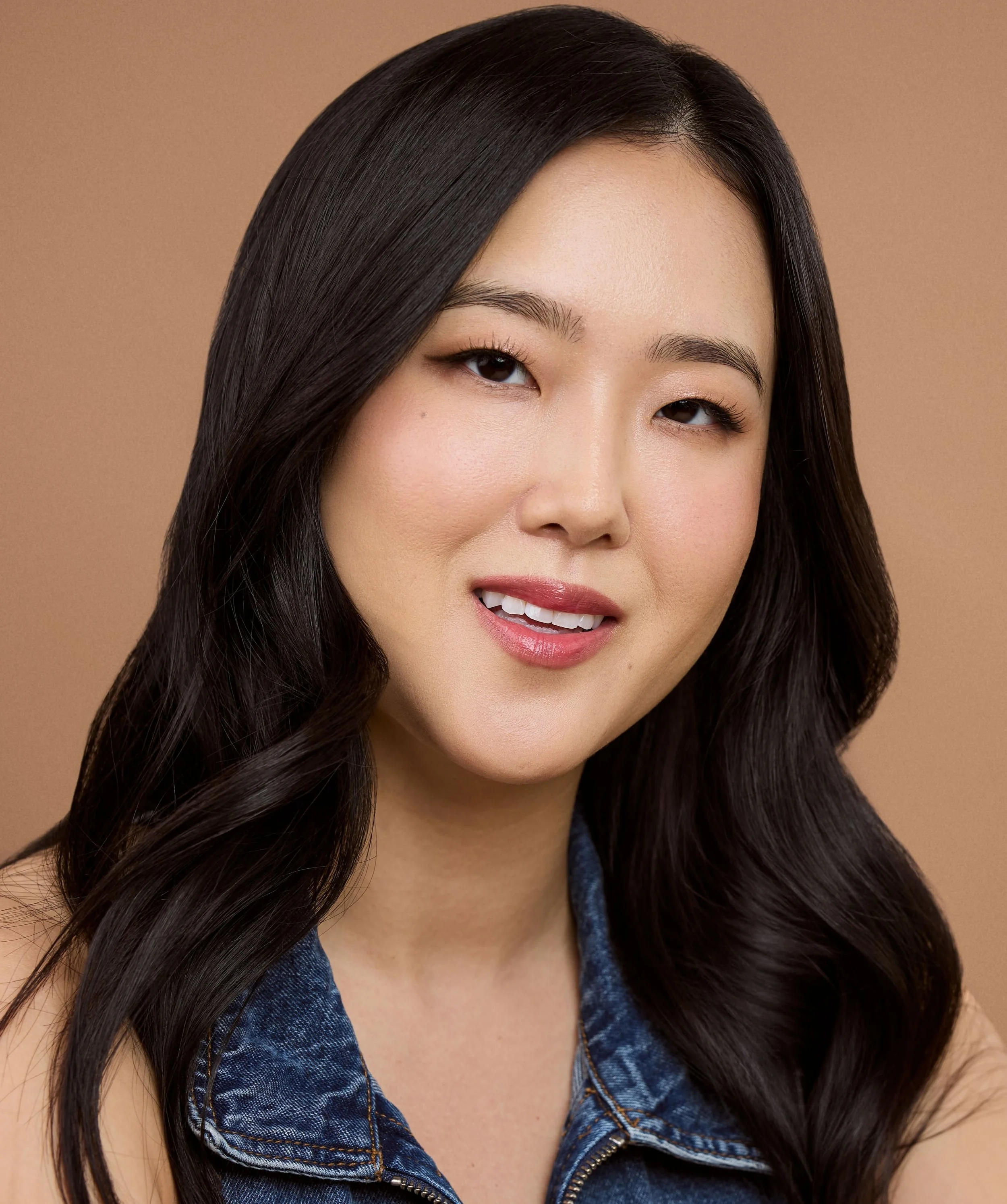 Close-up portrait of a woman with long dark hair, lightly smiling, wearing a denim top, against a beige background.