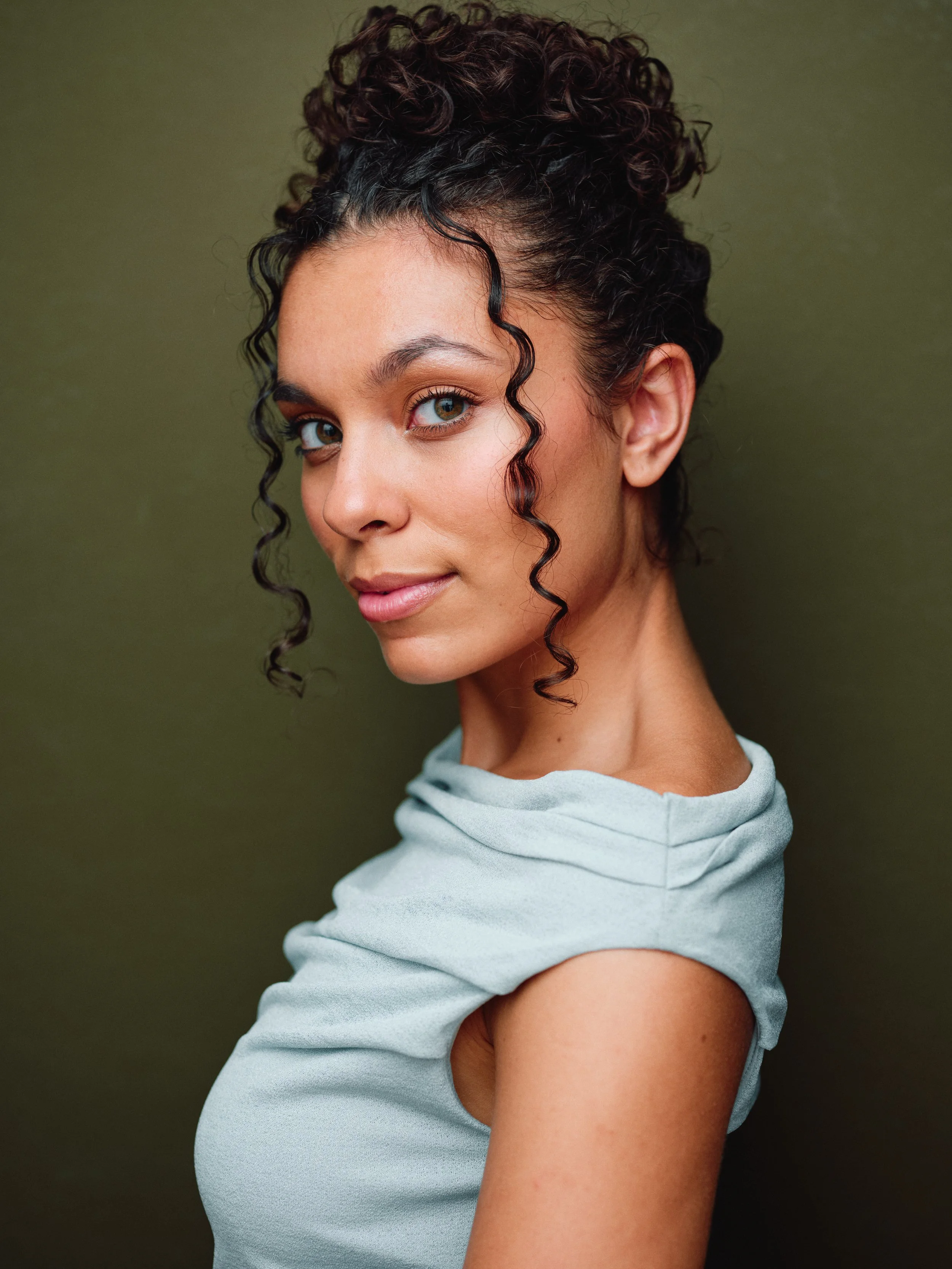 Close-up portrait of a young woman with curly dark hair, blue eyes, and light skin, wearing a light gray sleeveless top, against a plain olive-green background.