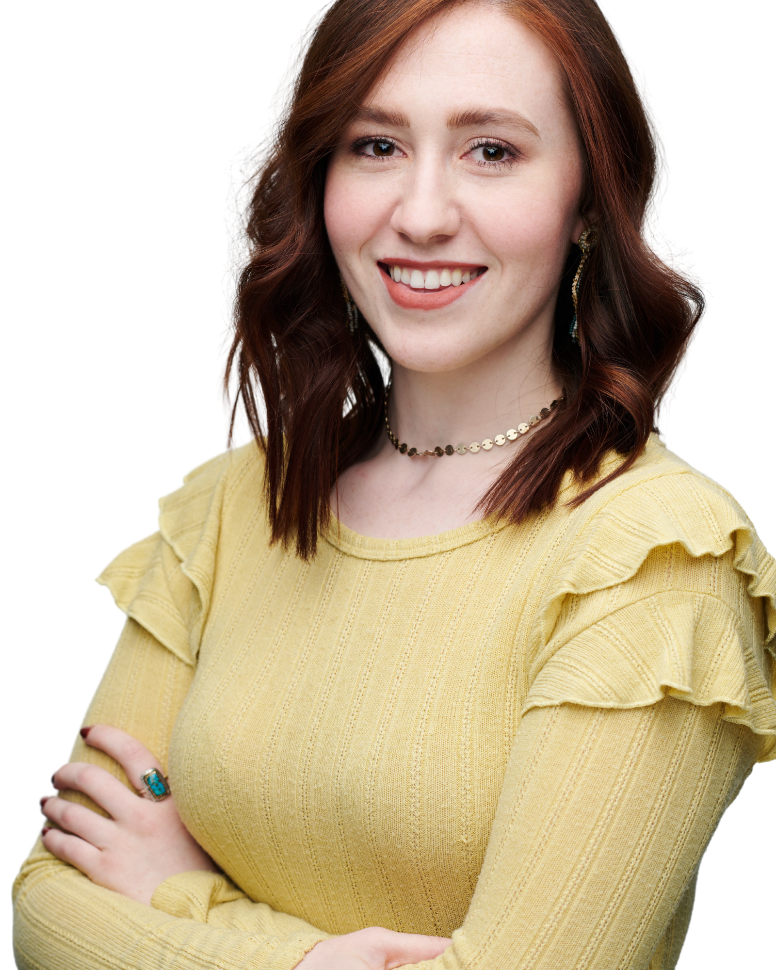 A young woman with red hair smiling, wearing a yellow top with ruffle details on the shoulders, layered necklaces, a ring, and earrings, with arms crossed.