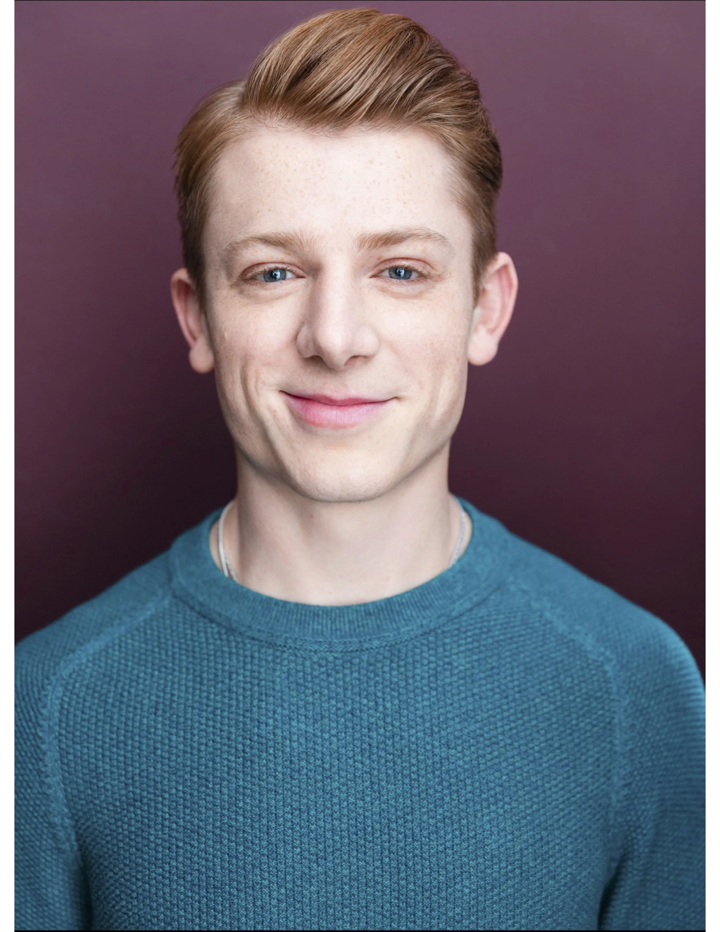 Close-up portrait of a young man with fair skin, red hair, and blue eyes, wearing a teal sweater, smiling softly against a purple background.