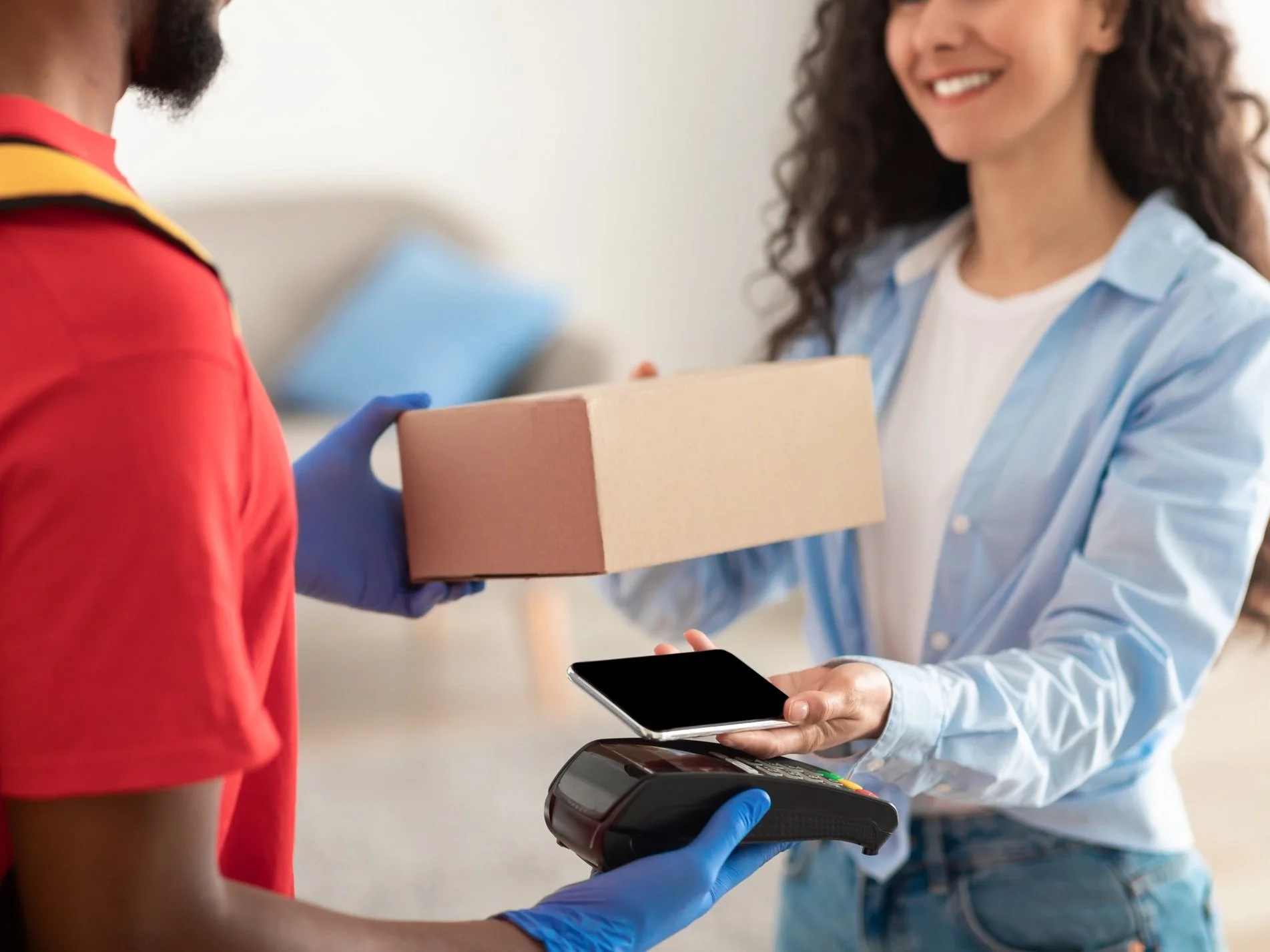 Une femme souriante remet un colis à une cliente, qui effectue un paiement avec son smartphone sans contact, à un point de vente.