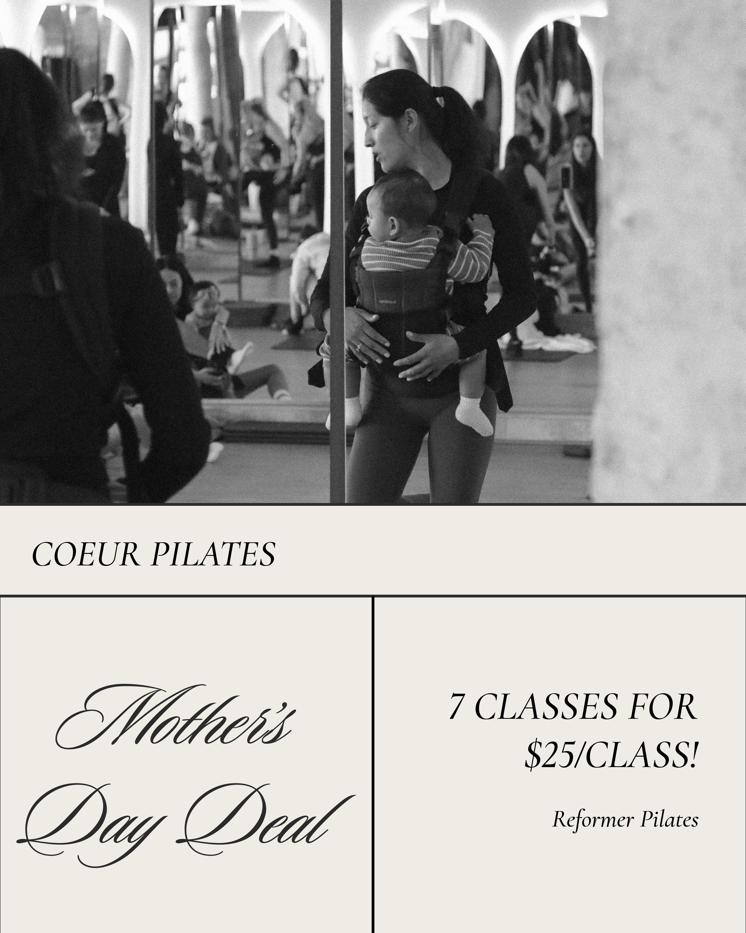 A woman holding a young child in a carrier during a Pilates class in a studio with mirrors, with several other people sitting and standing in the background.