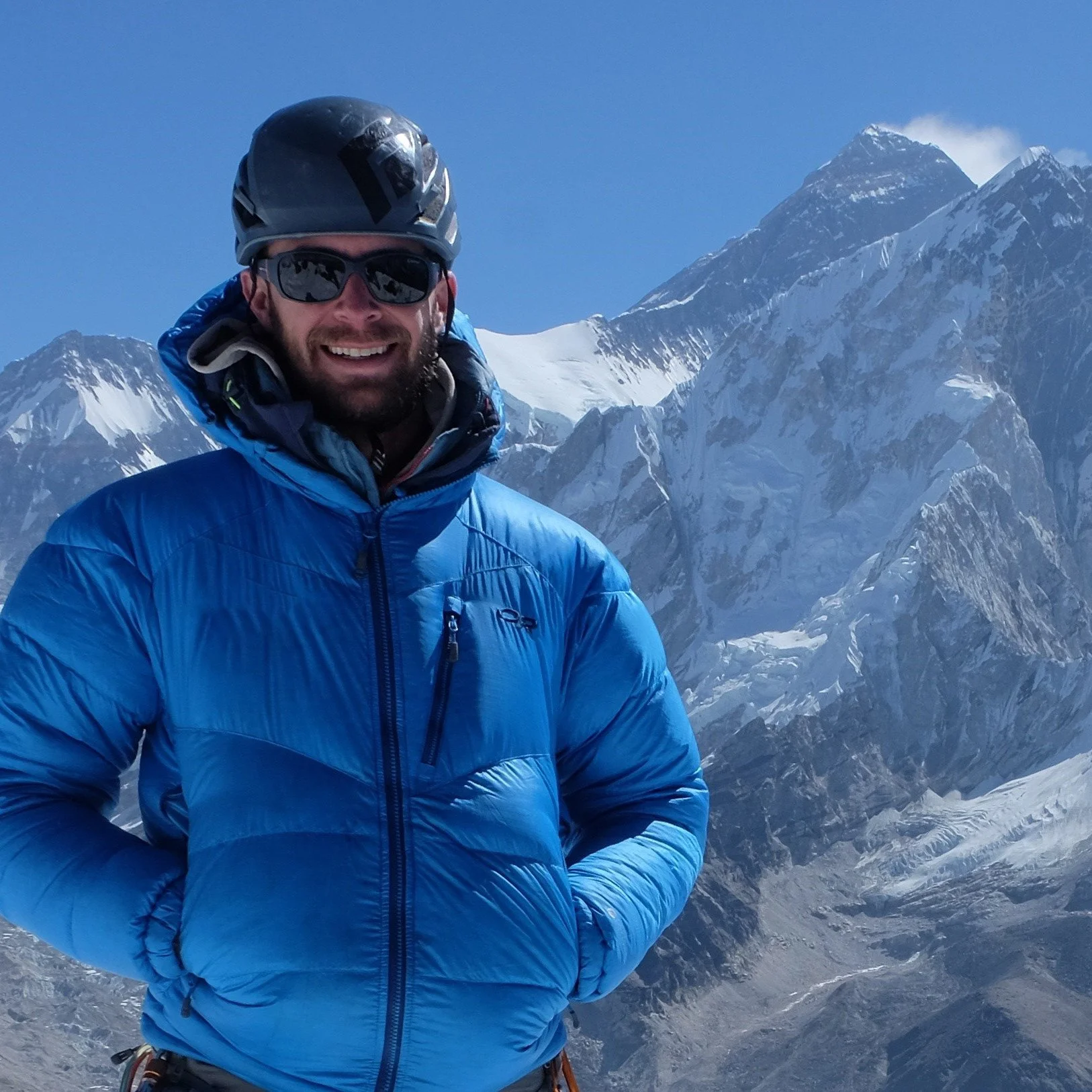 Rugged Luxury Guide, Dallas Glass, in winter gear and a helmet, standing in front of a mountain peak he guides clients on