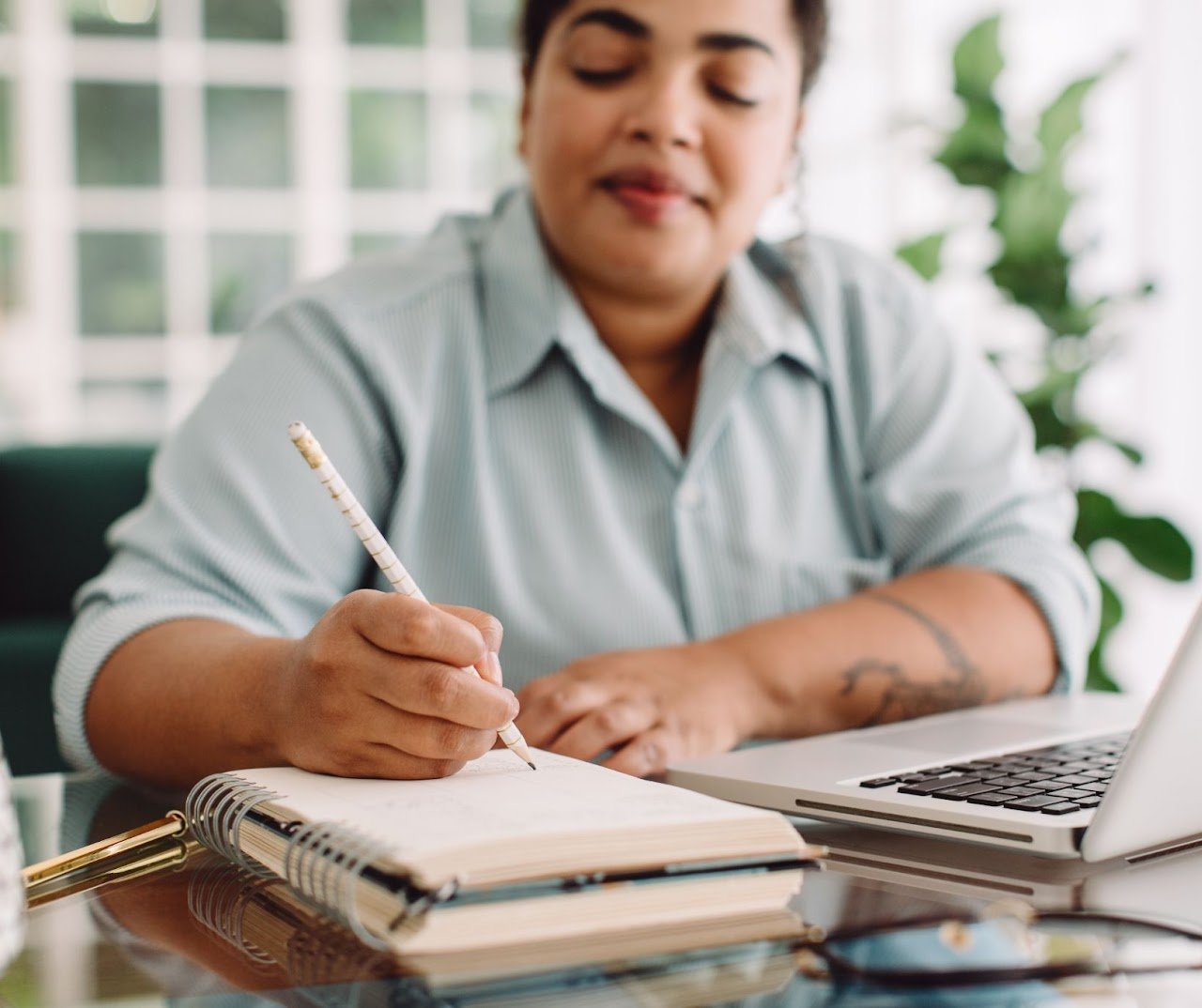 A person writing in a notebook at a desk with a laptop and other office supplies, in a well-lit room with a window and green plants in the background.
