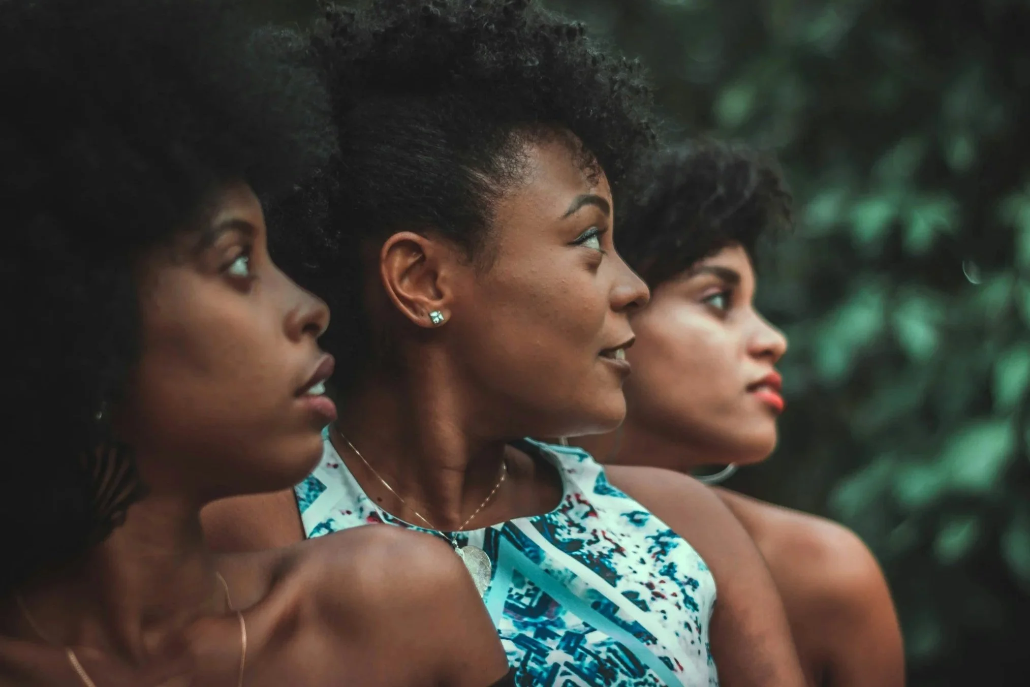 Three women with dark curly hair sitting closely together outdoors, facing to the right, with green foliage in the background.