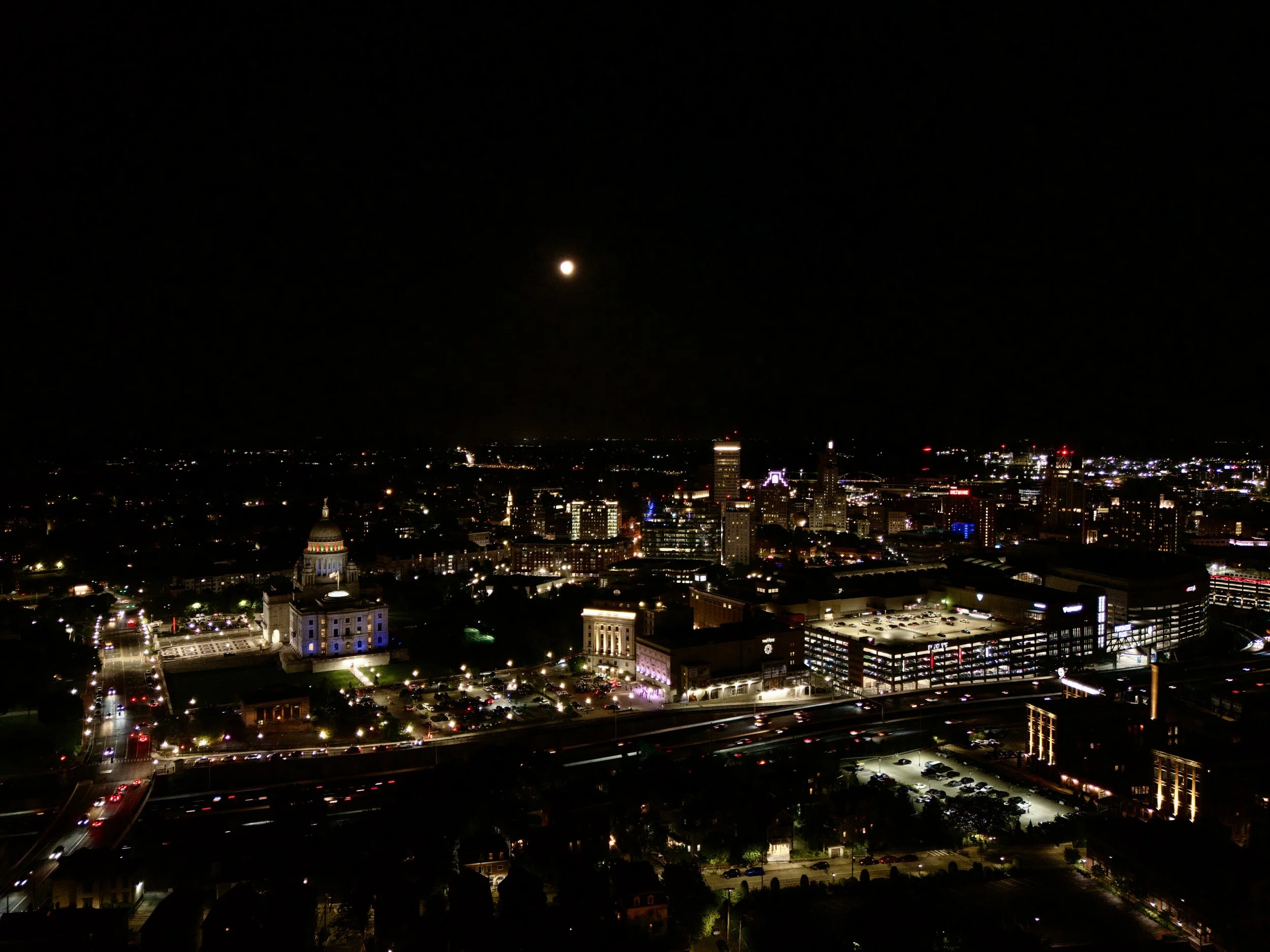 Nighttime aerial view of the Providence skyline with illuminated State House in the foreground, under a dark sky with a full moon.