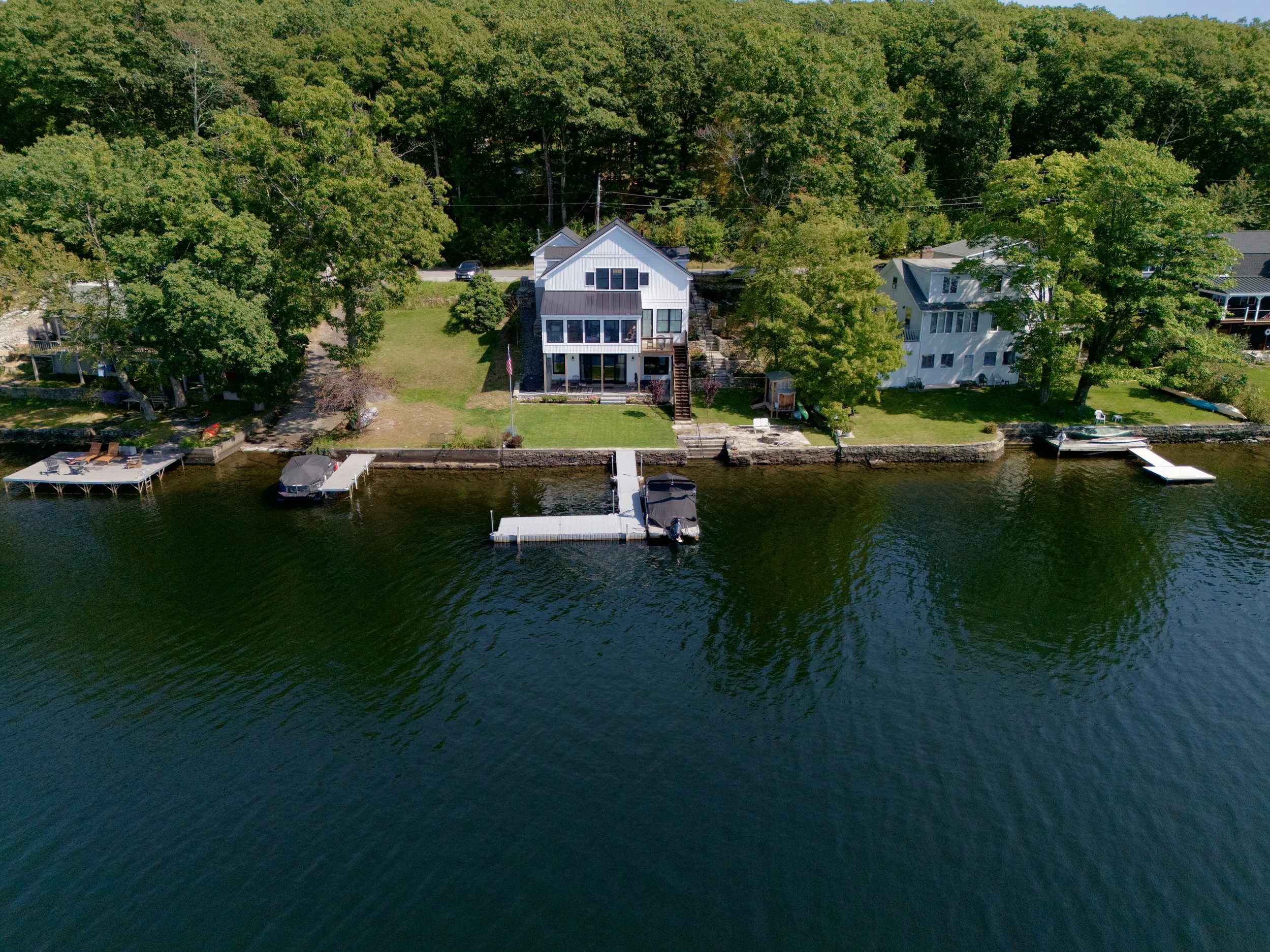 Altitude Drone Dynamics. Aerial view of a lakeside house with a large backyard, dock, and boat on the water. The house is white with a covered patio and stairs leading down to the dock with a boat anchored to it.