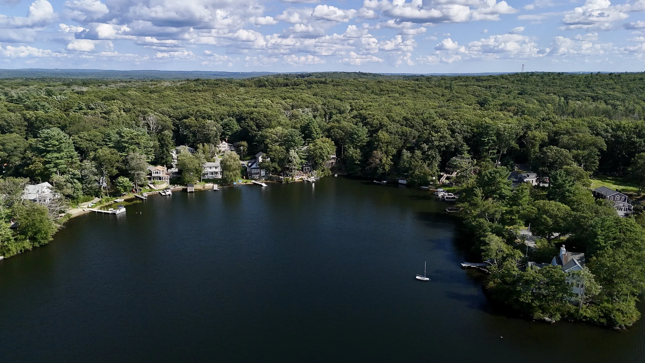 Altitude Drone Dynamics. Aerial view of a lakeside community property in Massachusetts with houses along the shoreline surrounded by dense green trees, under a partly cloudy sky.