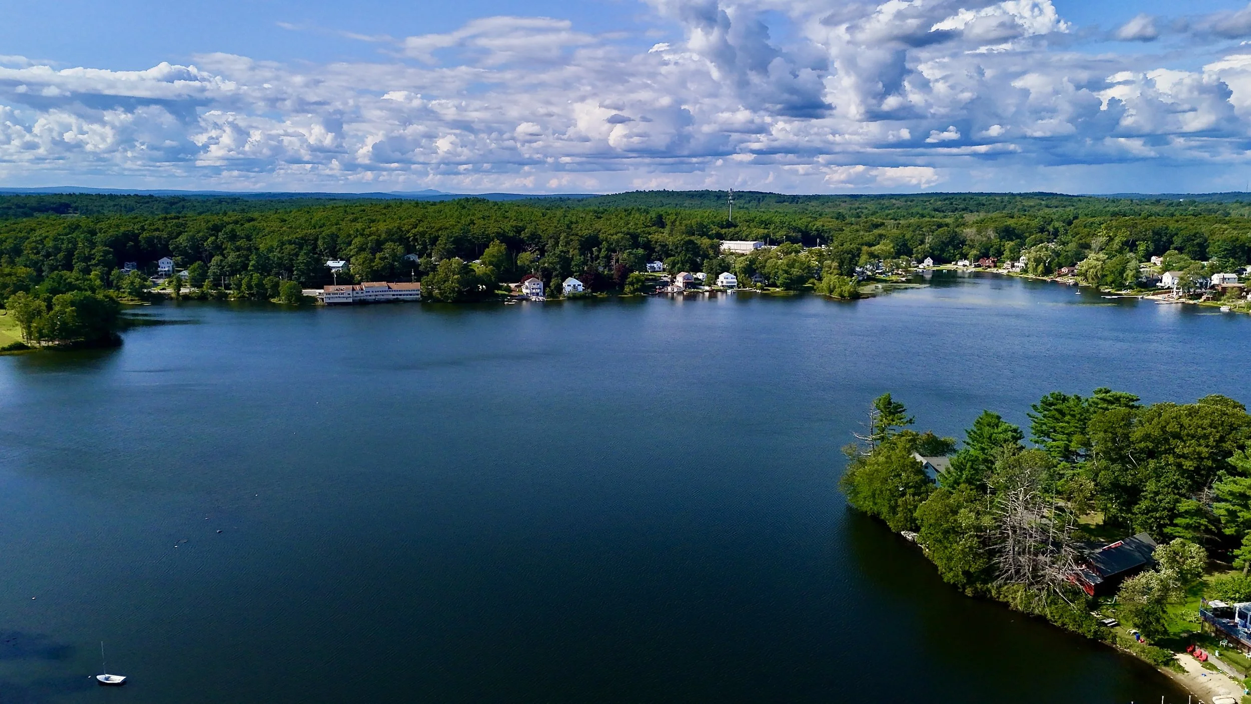 Altitude Drone Dynamics. Horizon view of a calm lake in New England, Central Massachusetts, Worcester surrounded by lush green trees and houses, with a partly cloudy sky overhead.